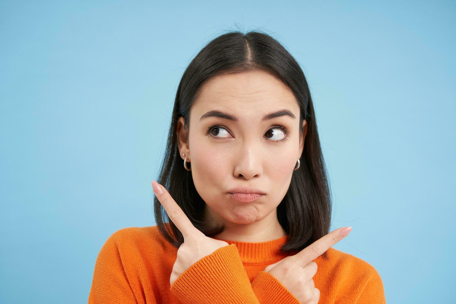 Close up of puzzled asian girl, points left and right, shows two choices, makes decision, stands over blue background photo