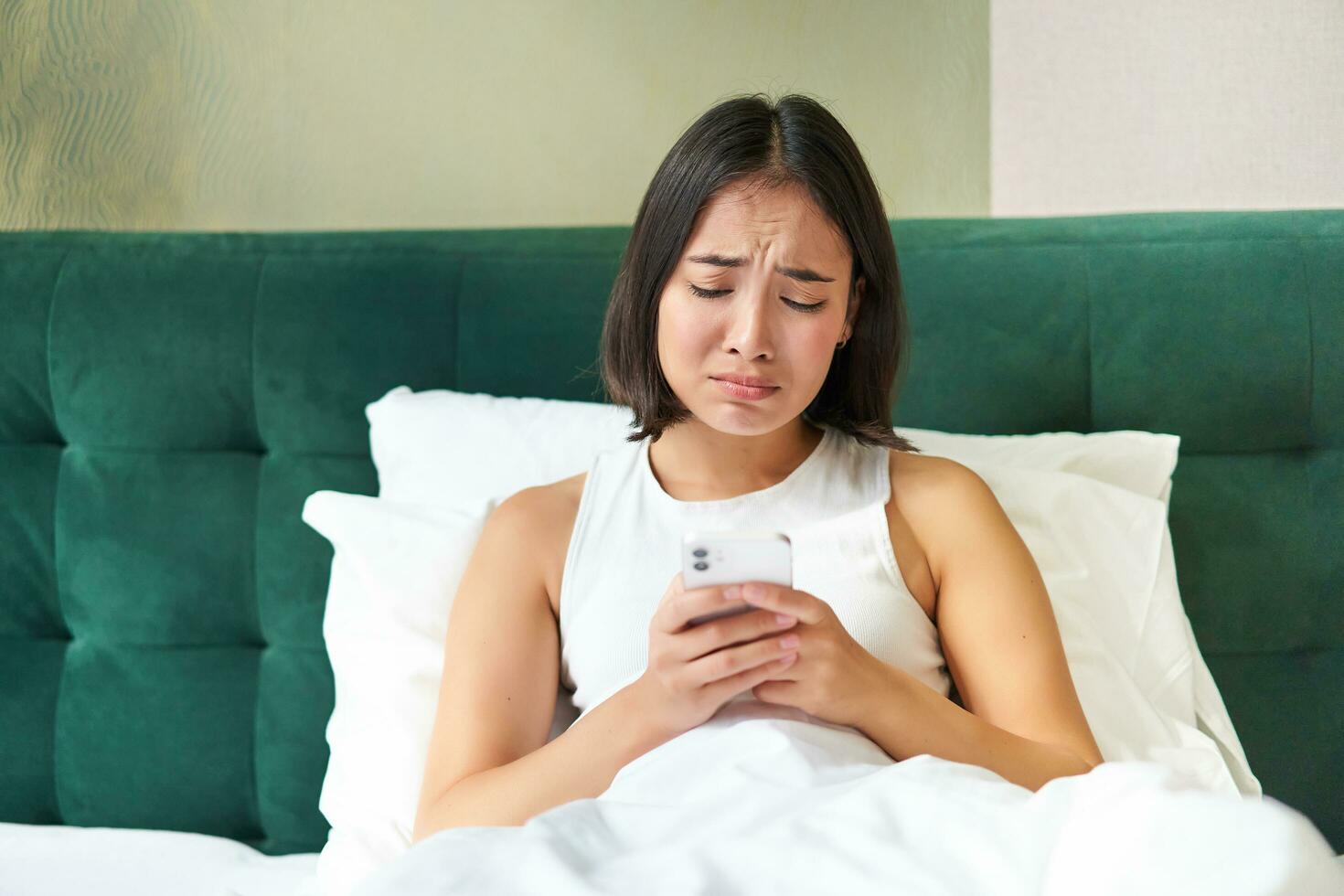 Disappointed asian girl, crying and whining, feeling upset, lying in bed on pillow, holding mobile phone, read upsetting bad news, waking up in awful mood photo