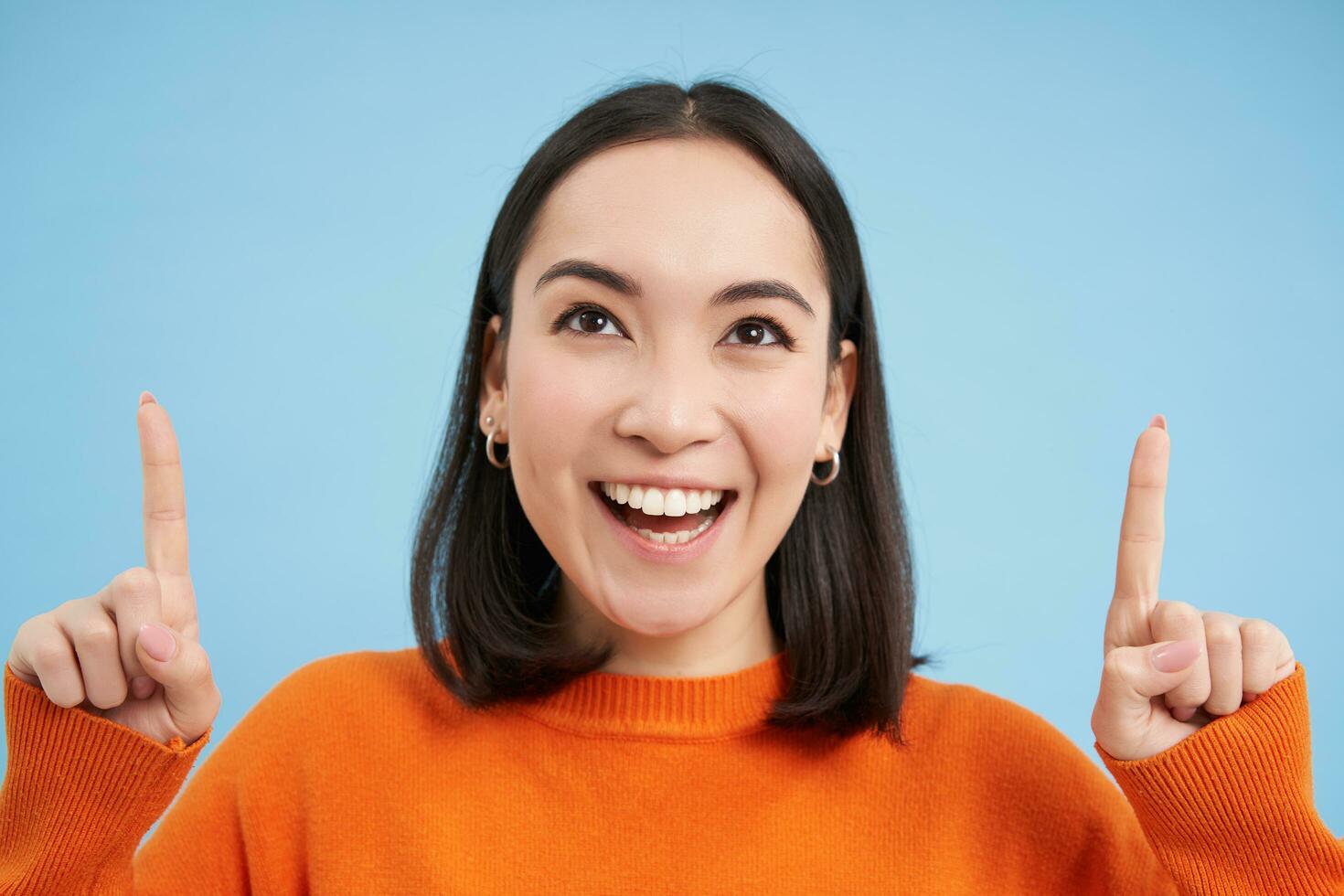 Beautiful asian girl points fingers up, shows banner on top, advertisement, stands over blue background photo