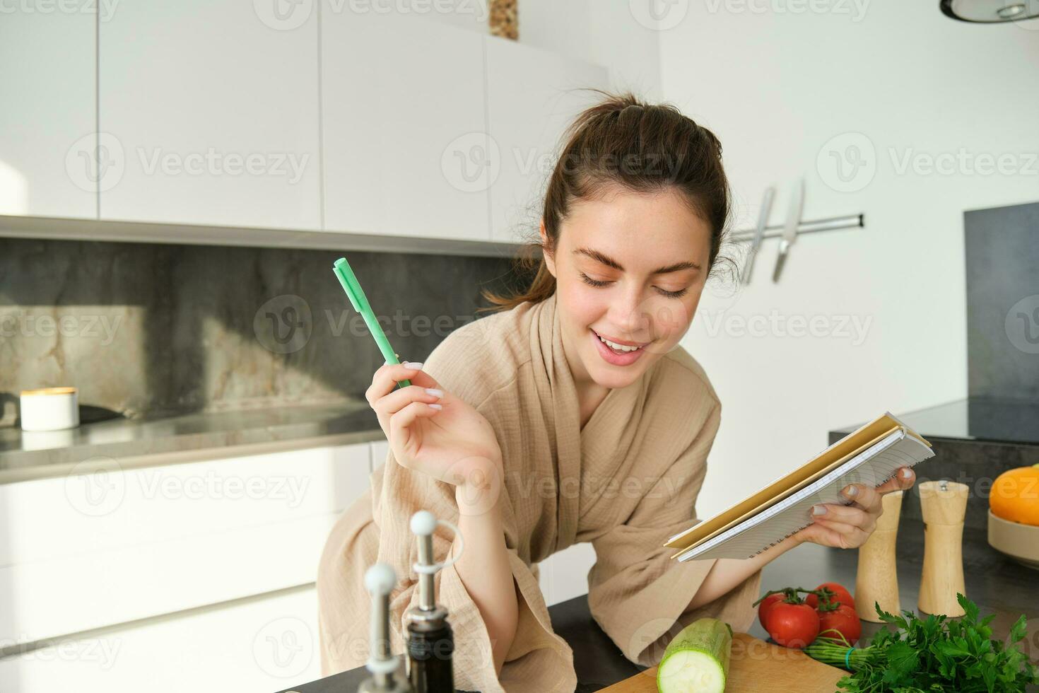 Portrait of woman checking grocery list, looking at vegetables, holding notebook, reading recipe while cooking meal in the kitchen, chopping tomatoes and zucchini photo