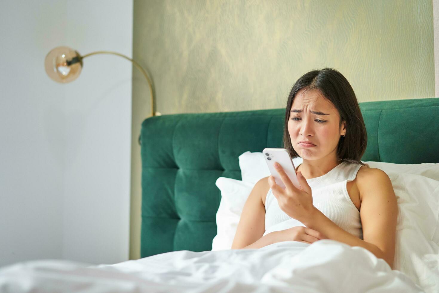 retrato de preocupado asiático mujer participación móvil teléfono, recibir malo teléfono llamar, mirando preocupado y decepcionado, teniendo difícil teléfono conversacion mientras acostado en cama foto