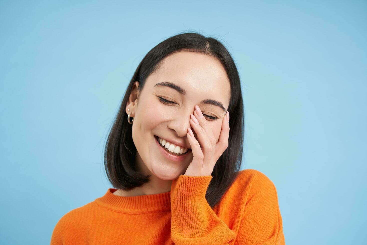 Close up of beatiful brunette woman, laughing and smiling, touches her face, expresses pure joy, stands over blue background photo