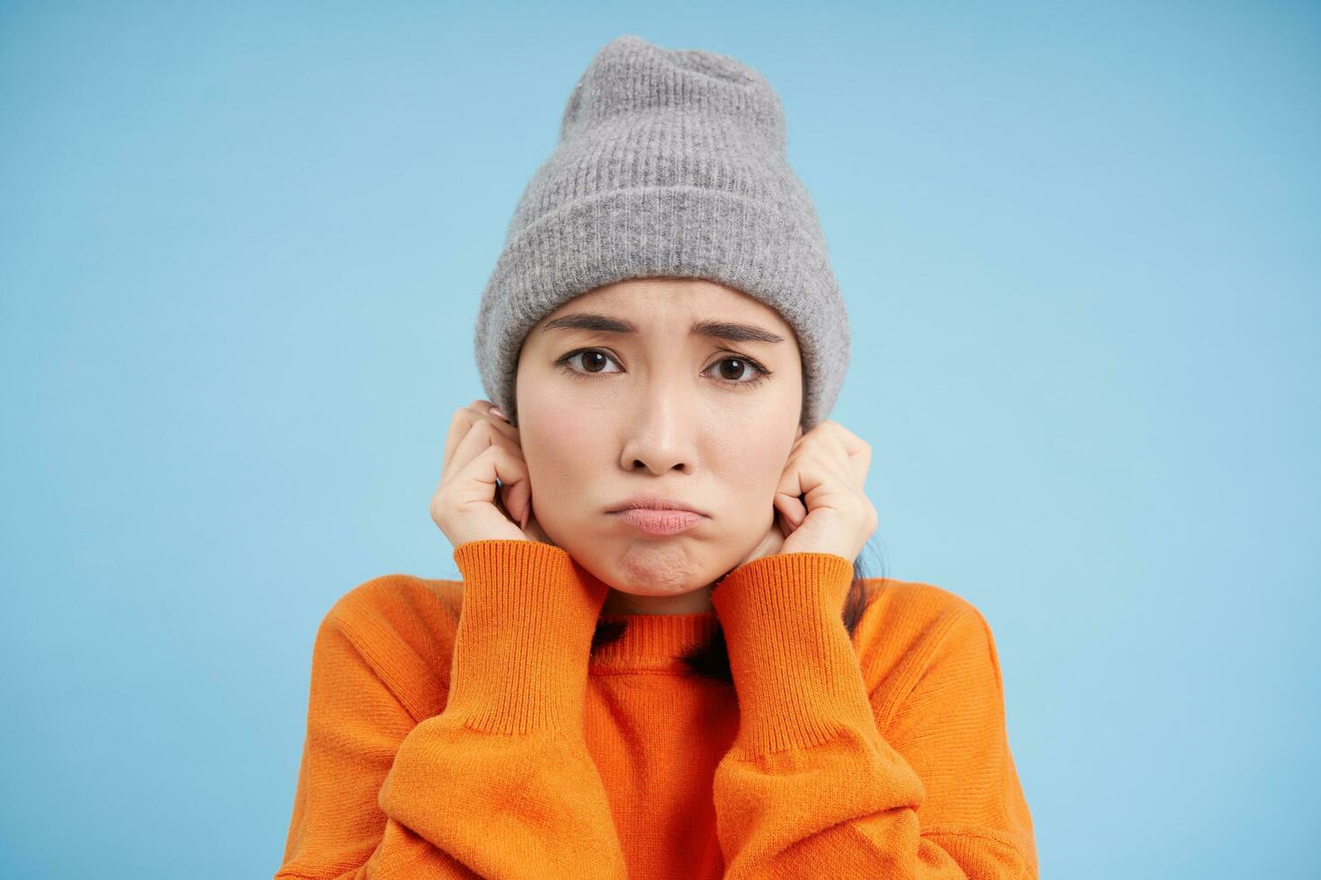 cerca arriba retrato de coreano niña siente frío, batidos y se congela, pone en gorro y se enfurruña con triste rostro, soportes terminado azul antecedentes foto