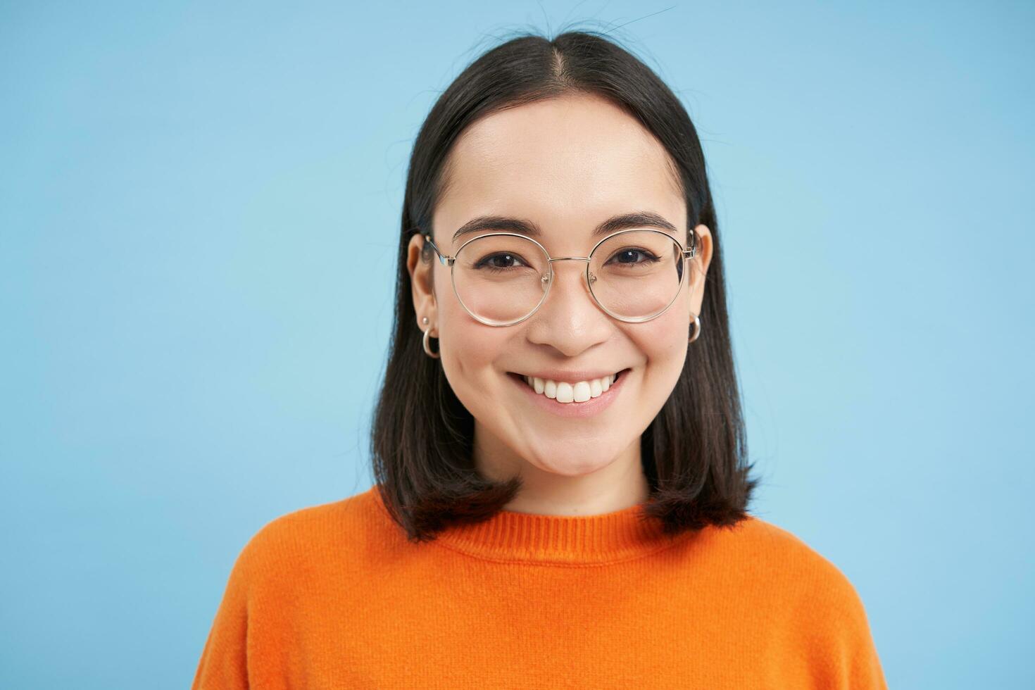 Beauty and skincare. Close up portrait of happy smiling japanese woman, touches her clear, glowing skin, natural healthy face, standing over blue background photo