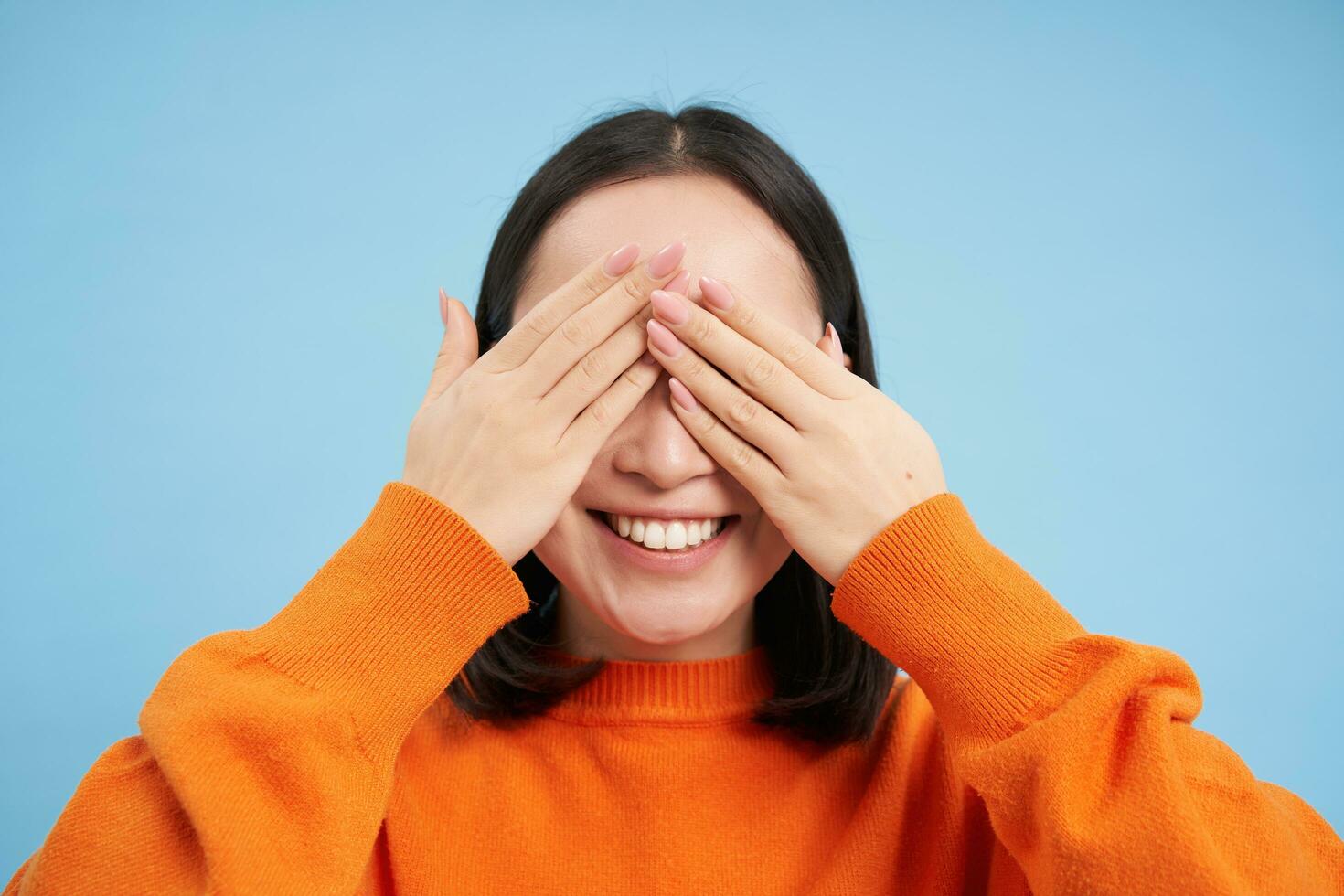 Close up of asian woman shuts her eyes and smiles blindfolded, waits for surprise, stands over blue background photo