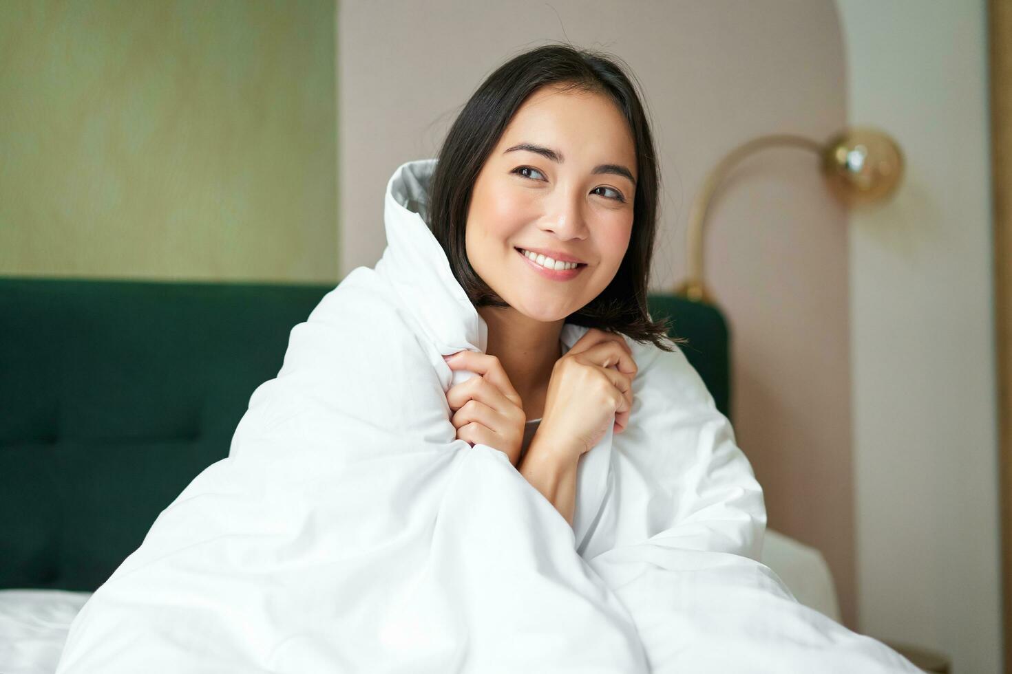 Beautiful romantic woman covers herself with warm duvet, enjoys sleeping and staying in bed, waking up happy in morning, sitting in bedroom photo