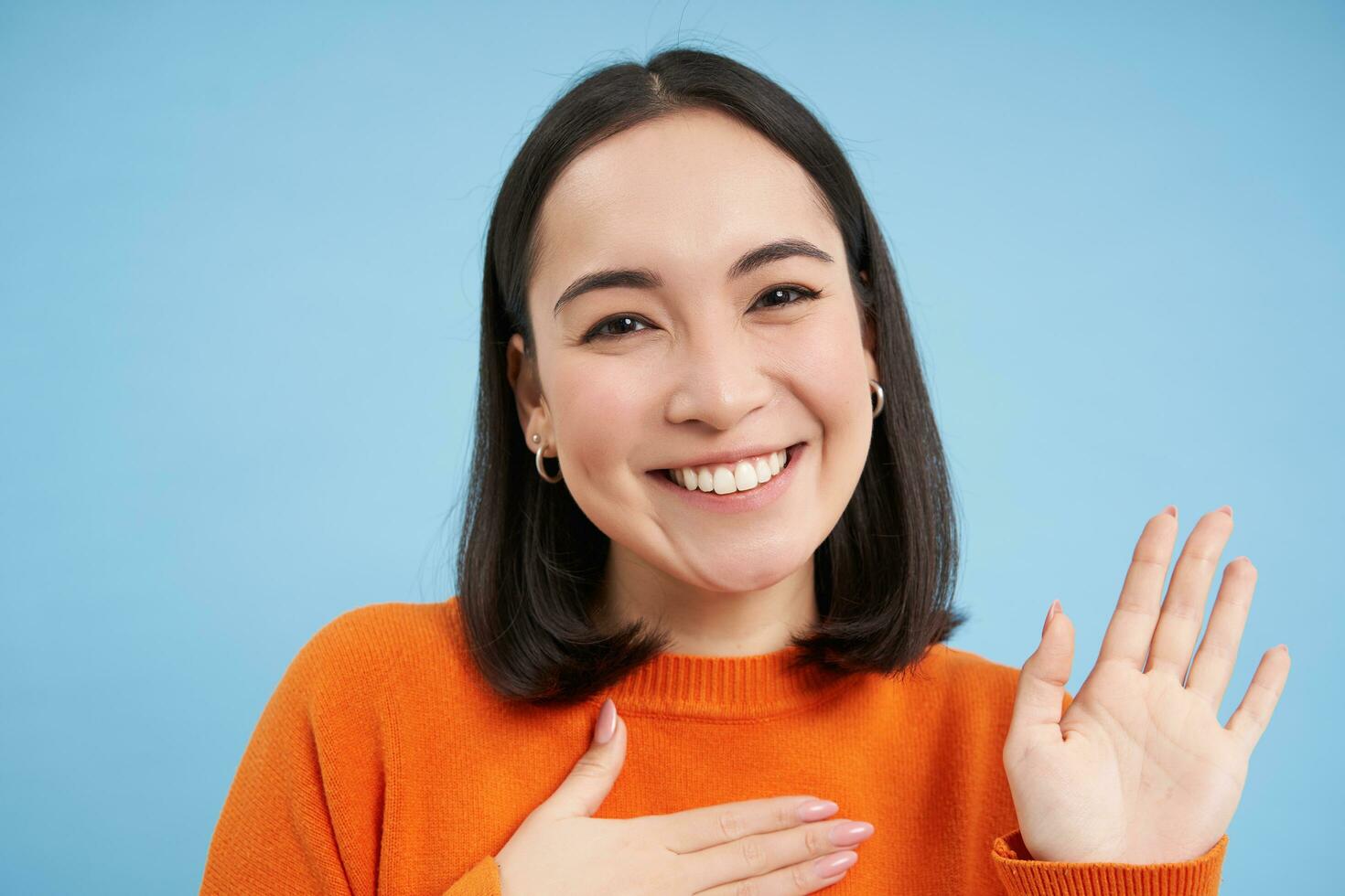 Close up of friendly smiling japanese woman, raises one arm, introduce herself, greets, stands over blue background photo