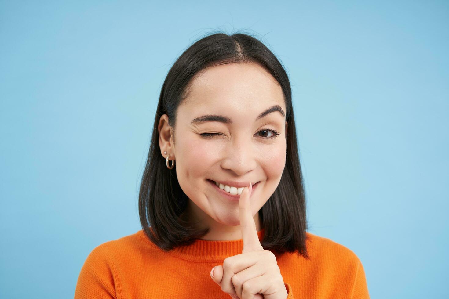 Close up of smiling asian girl, winks at you, tells secret, holds finger on lips, stands over blue studio background photo