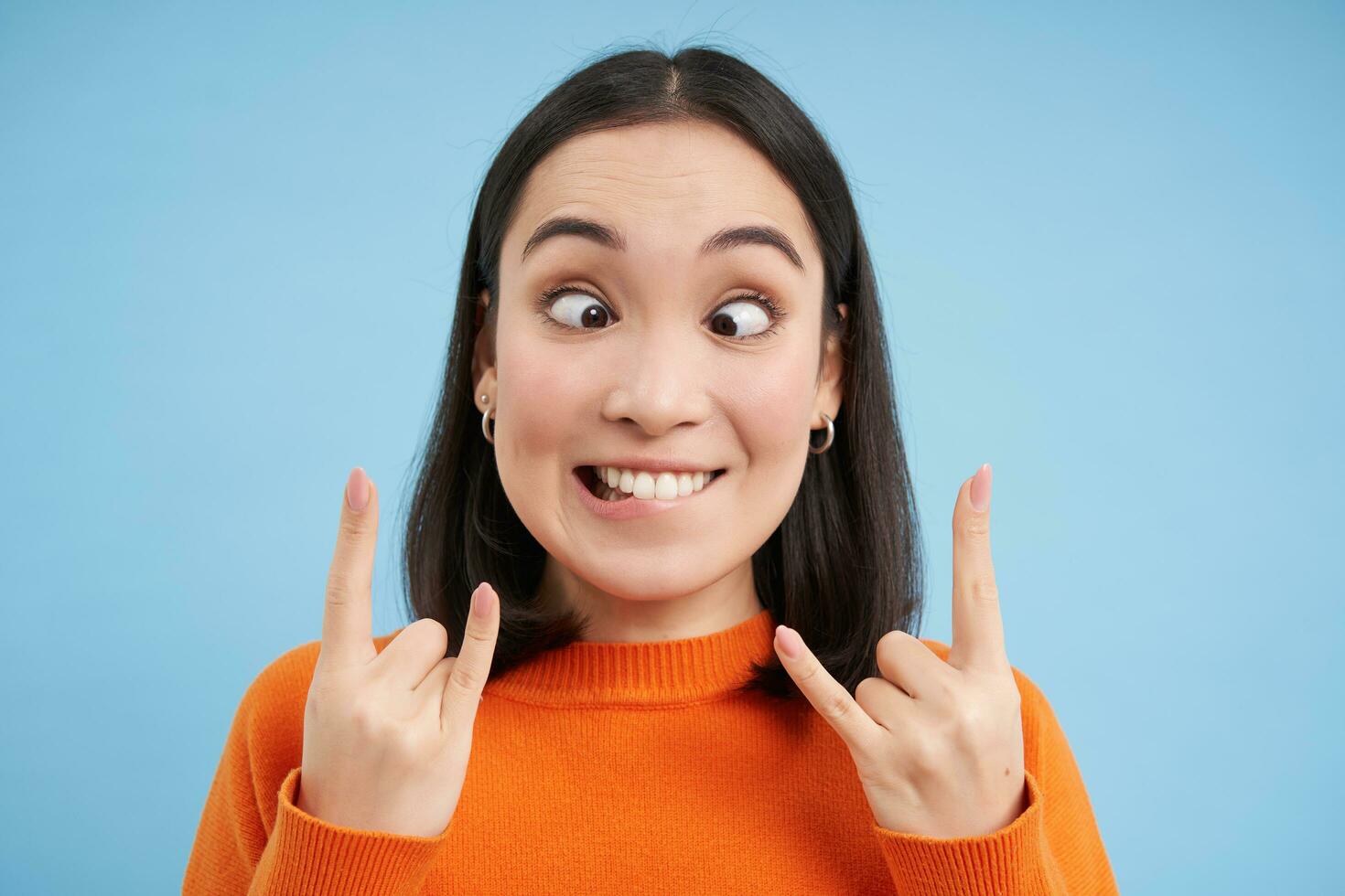 Close up of happy korean woman, shows rock n roll, heavy metal gesture, having fun, standing over blue studio background photo