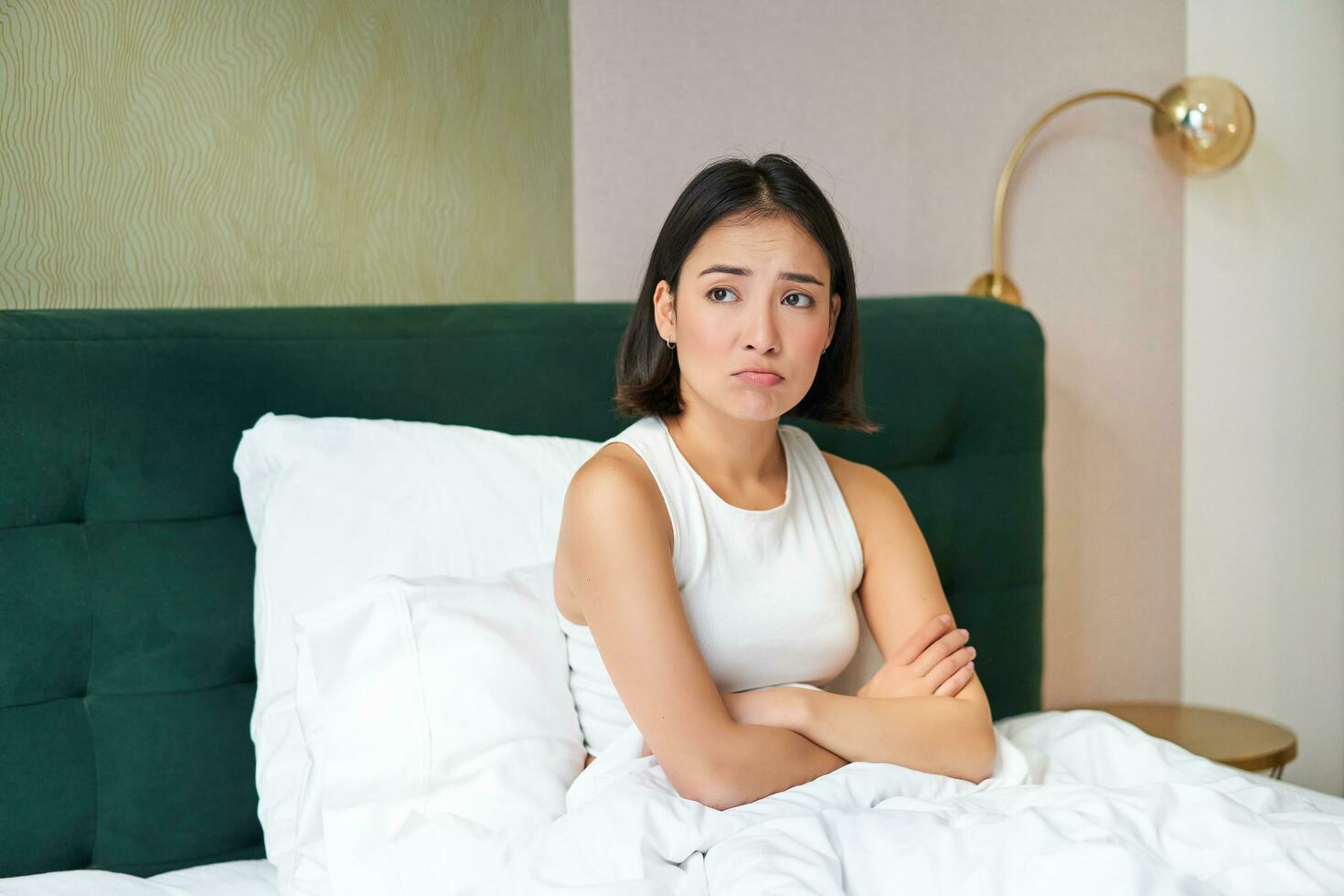 Portrait of asian girl in bed, looking complicated and worried, cross arms on chest, frowning with uneasy, thinking face expression photo