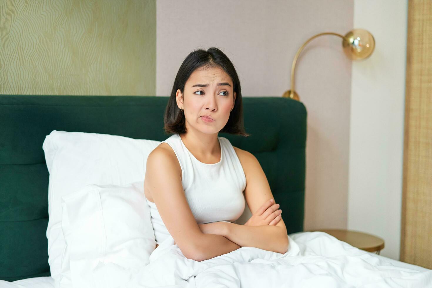 Portrait of asian girl in bed, looking complicated and worried, cross arms on chest, frowning with uneasy, thinking face expression photo