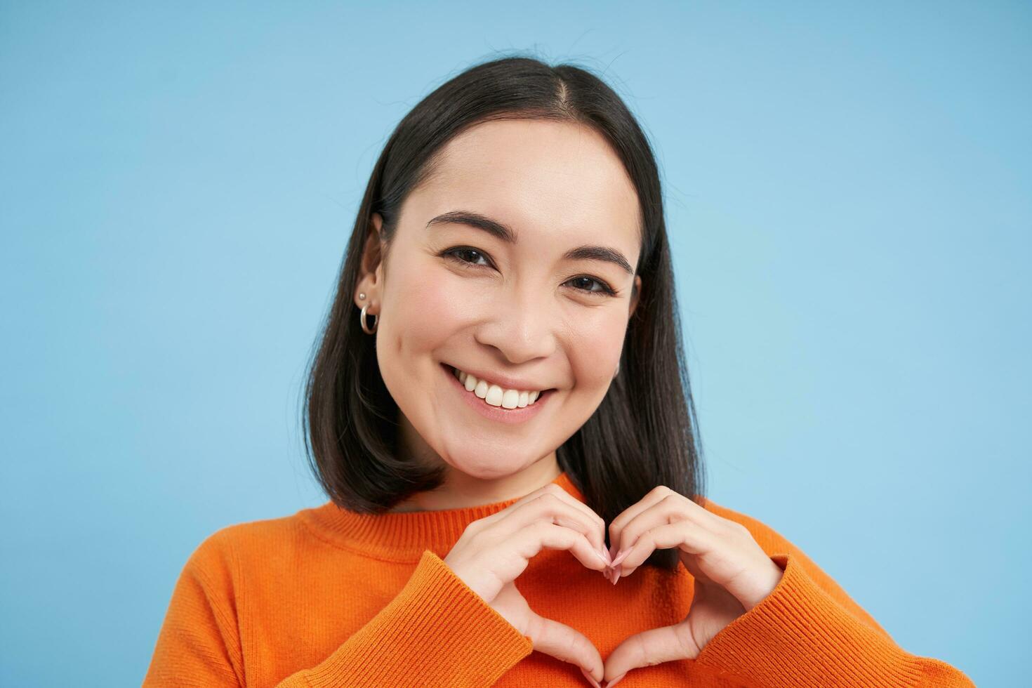 Close up portrait of lovely asian woman, shows heart sign and smiles with care and tenderness, stands over blue background photo