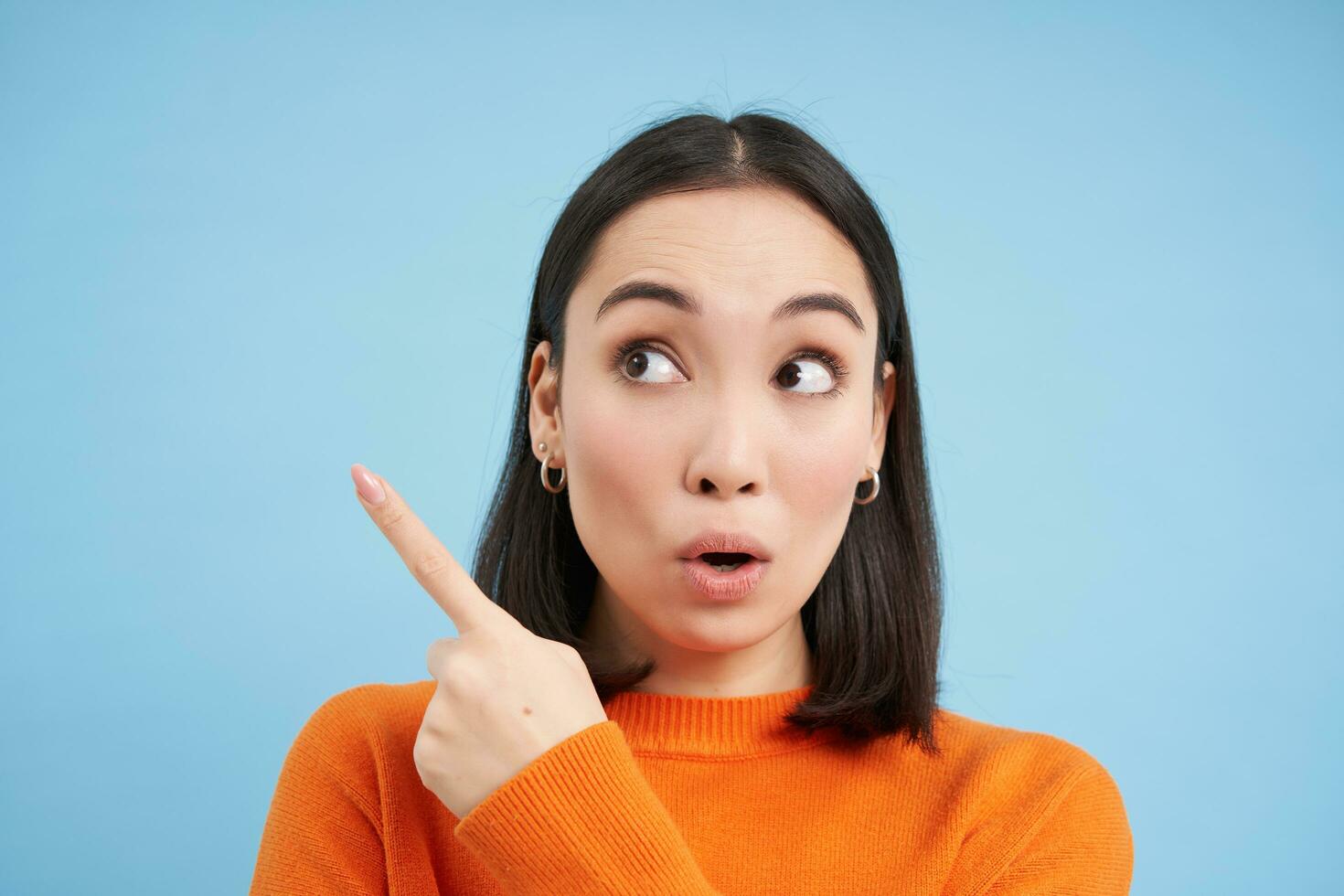 Close up portrait of asian girl, looks surprised, points at smth interesting, amazed by advertisement, blue studio background photo