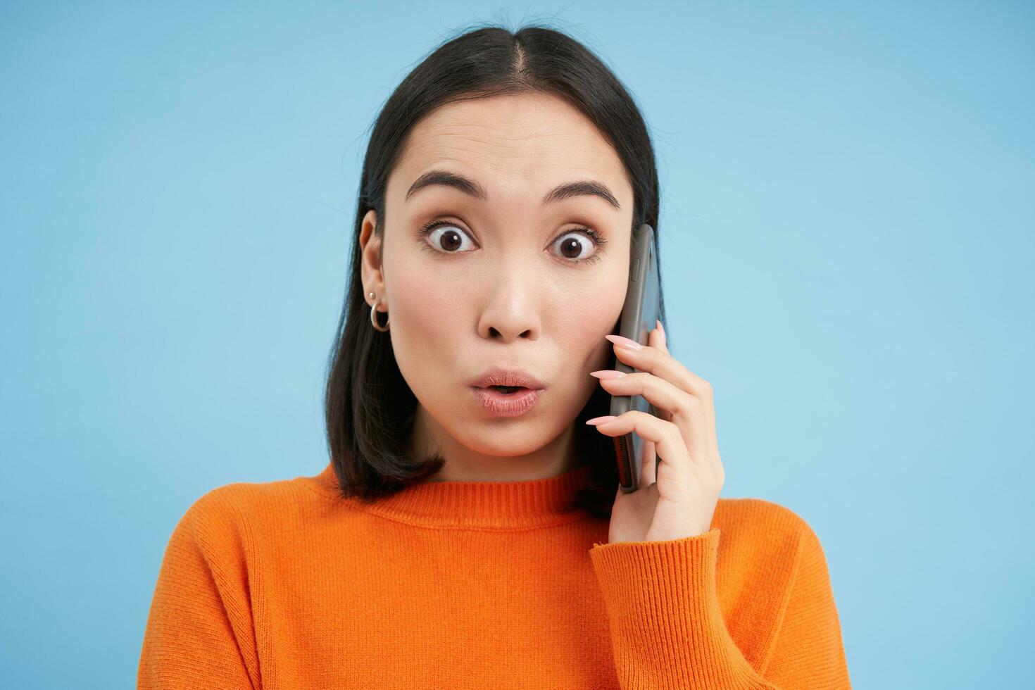 Technology concept. Asian girl answers phone call and looks surprised at camera, stands over blue background photo