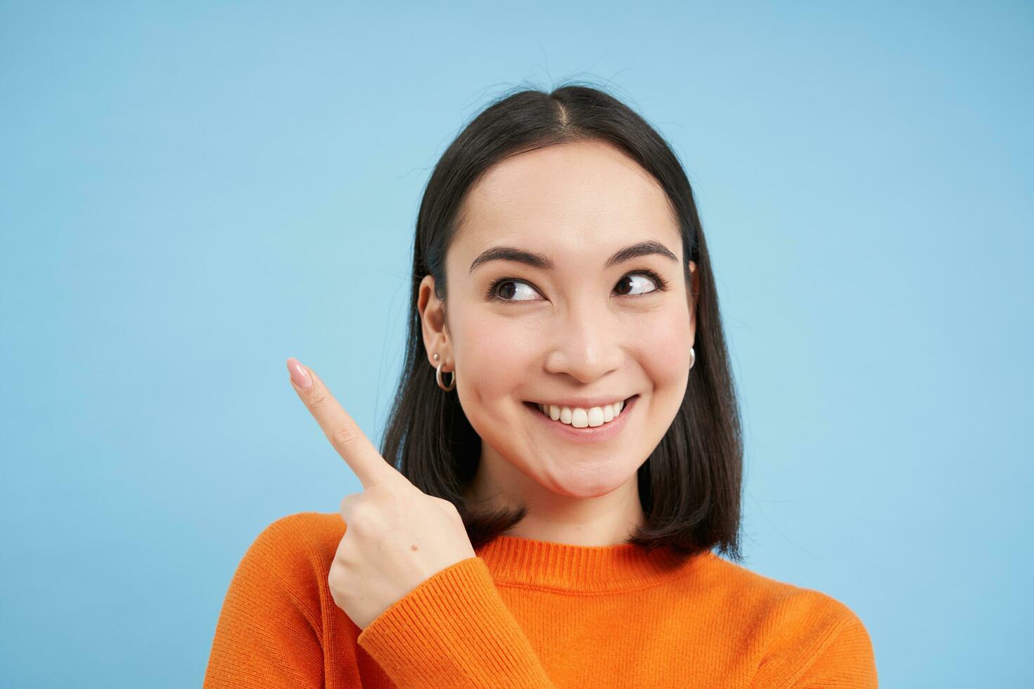 cerca arriba Disparo de sonriente asiático chica, puntos dedo a Superior izquierda esquina, muestra bandera, demuestra anuncio publicitario, azul antecedentes foto