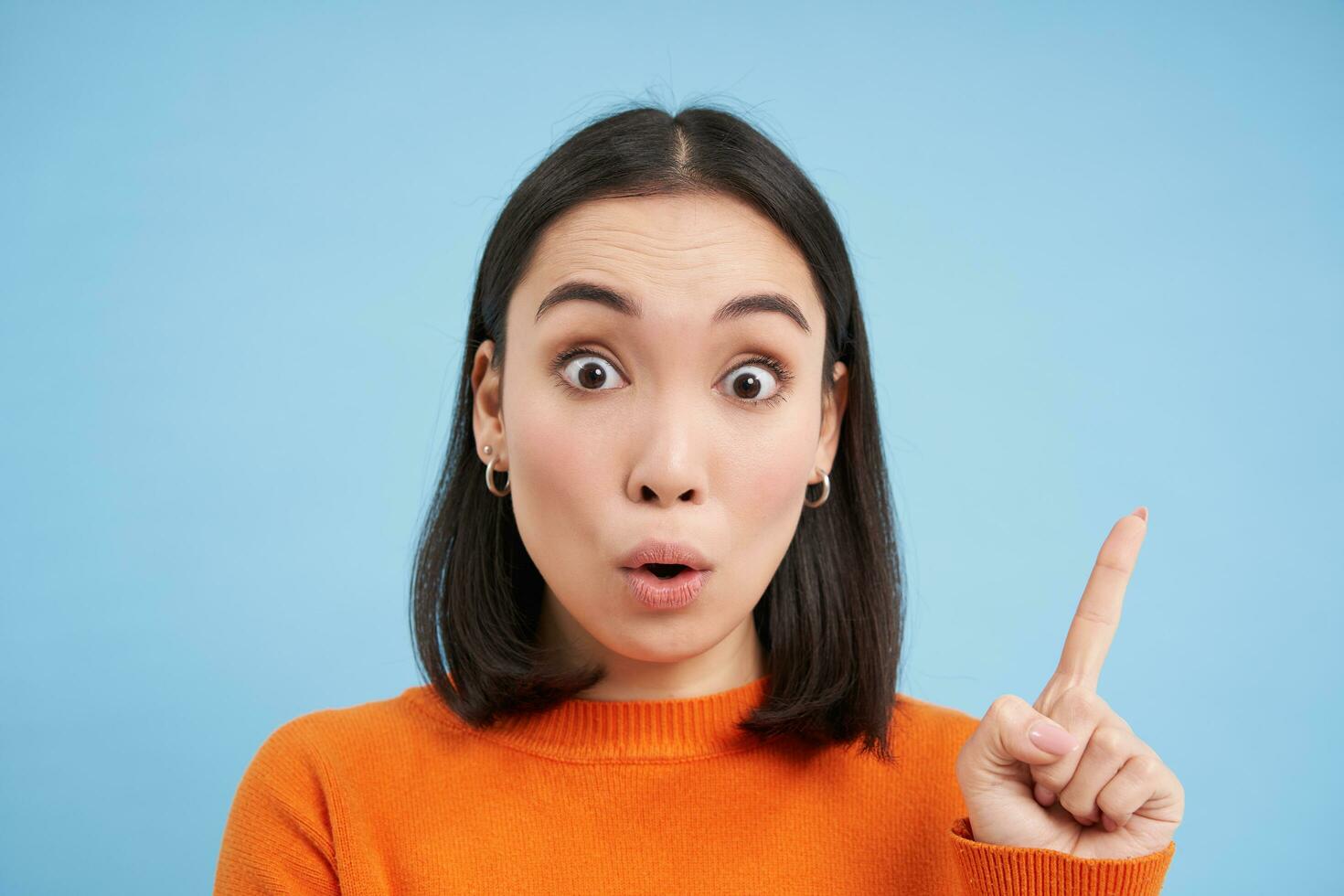 Close up portrait of asian girl, looks surprised, points at smth interesting, amazed by advertisement, blue studio background photo
