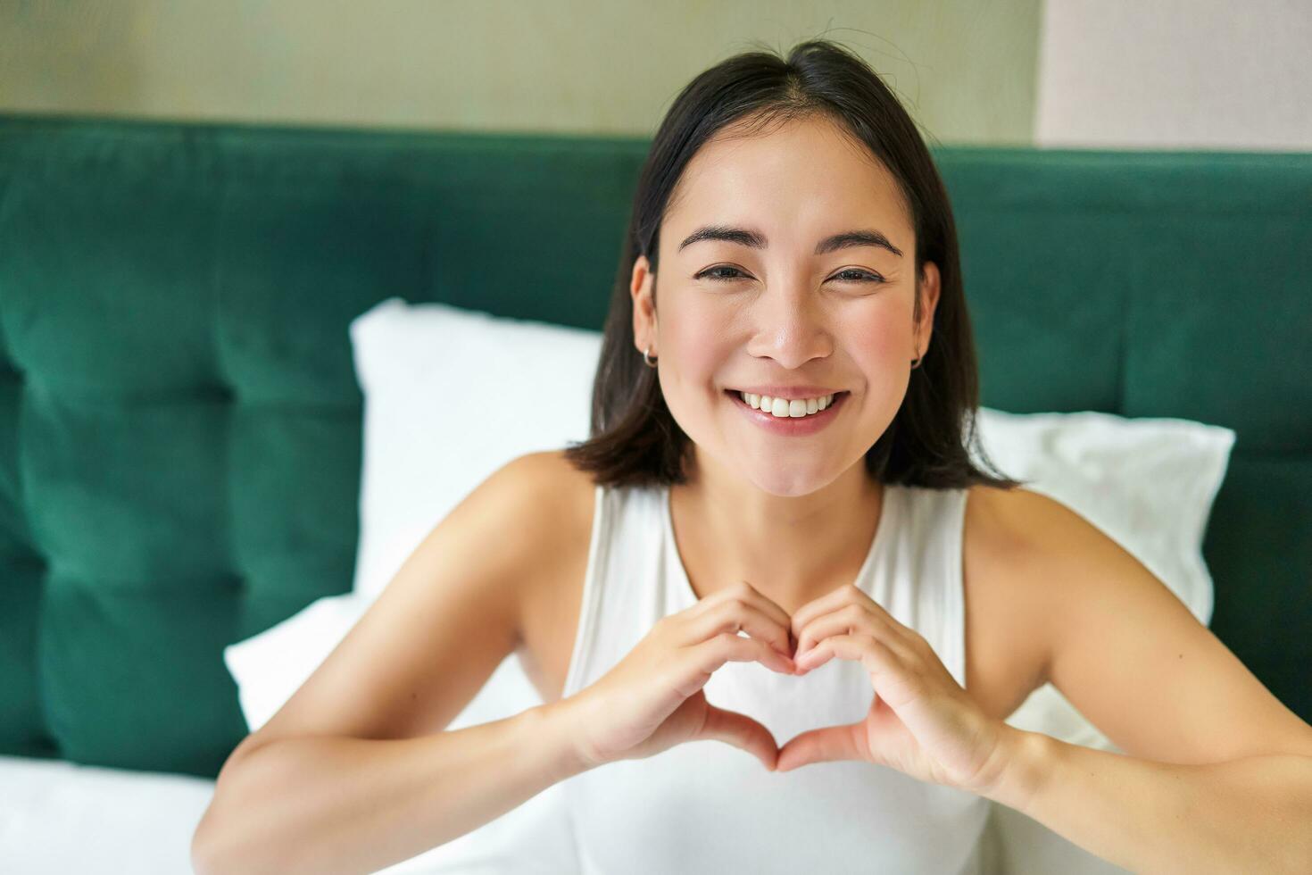 Cute asian girl shows heart, love sign, lying in bed and smiling, enjoying mornings, lazy days in bedroom photo