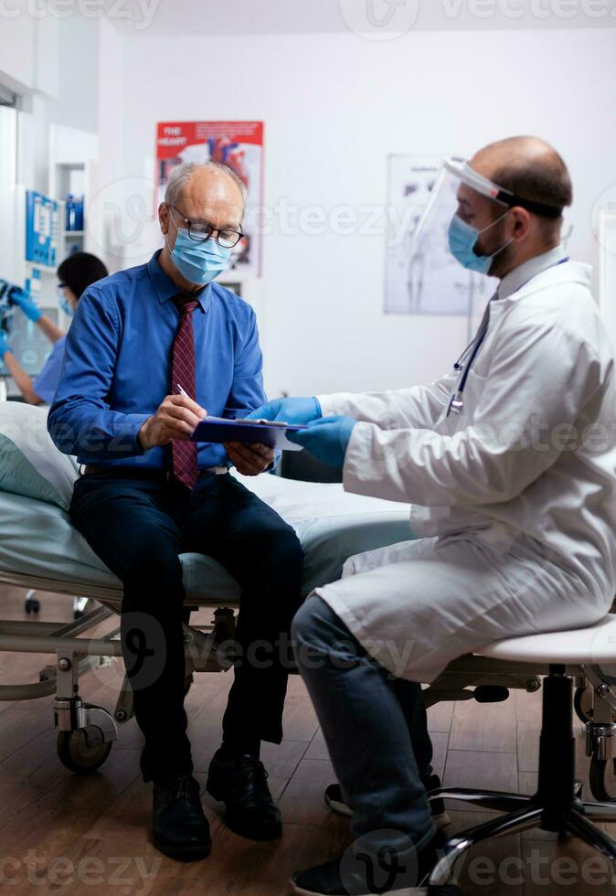 Senior patient wearing face mask agasint covid19 signs medical test results in hospital examination room with doctor sitting on bed. Healthcare medical physician consultation during global crisis. photo