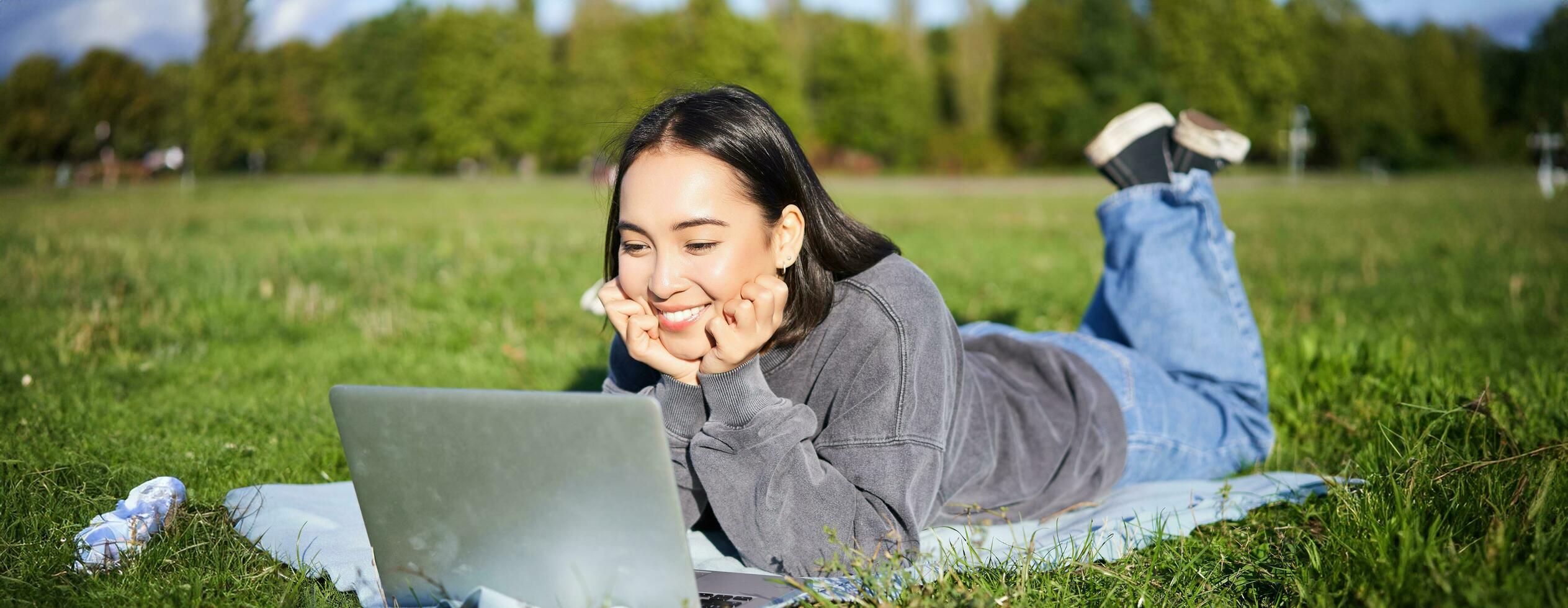 Smiling asian girl lying in park on grass, watching movie or video on laptop, looking at screen with interest photo
