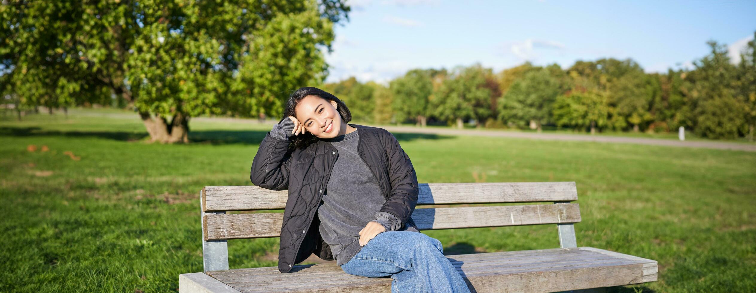 Smiling young asian woman in outdoor jacket, sitting on bench in green sunny park, resting alone, relaxing on fresh air photo