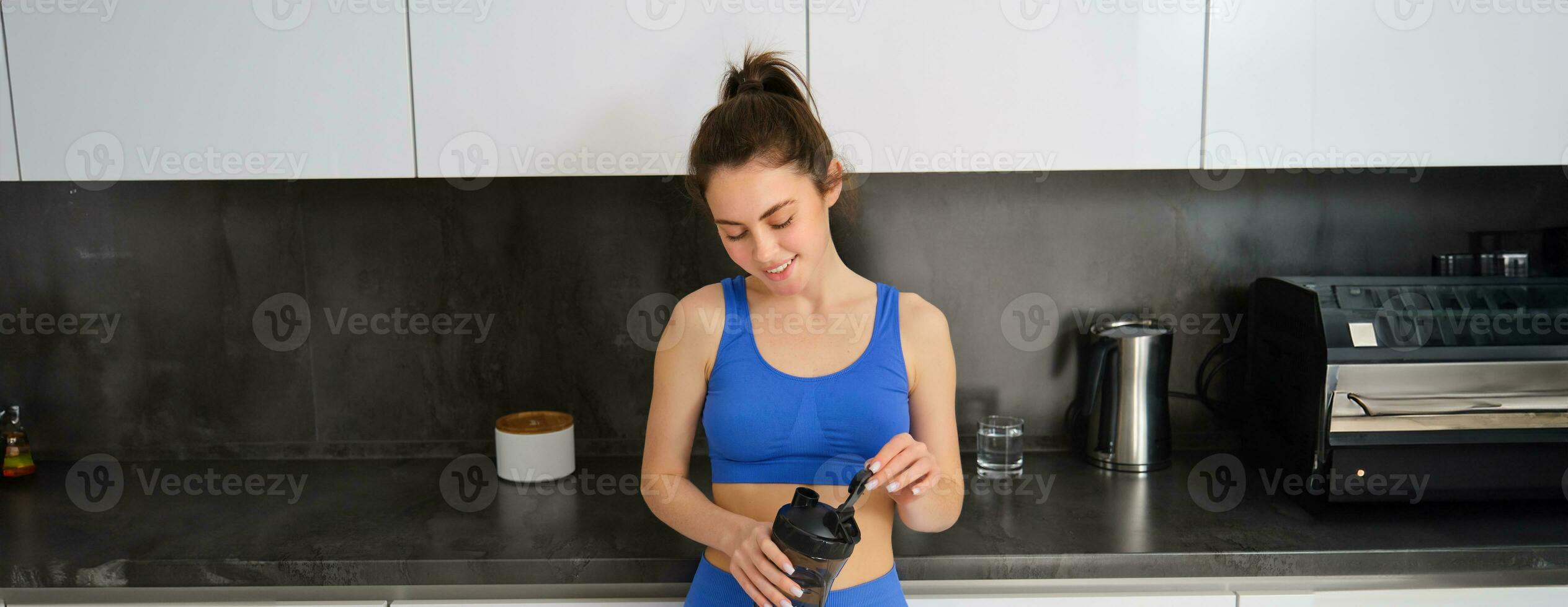 imagen de joven mujer preparando dietético suplemento, proteína en su criba vibradora botella, vistiendo aptitud ropa, en pie en cocina foto