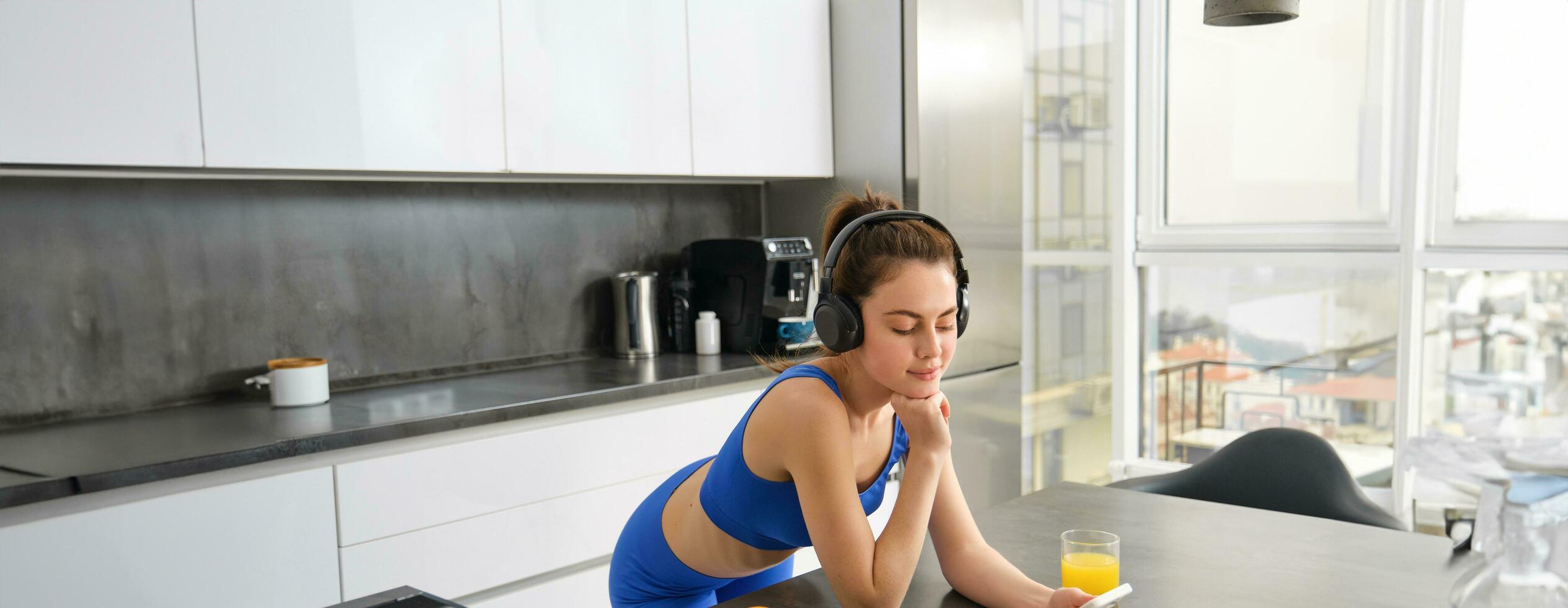 Portrait of young modern woman, beautiful girl in fitness clothes, standing in kitchen, watching yoga video class on smartphone, listening in wireless headphones, drinking juice photo