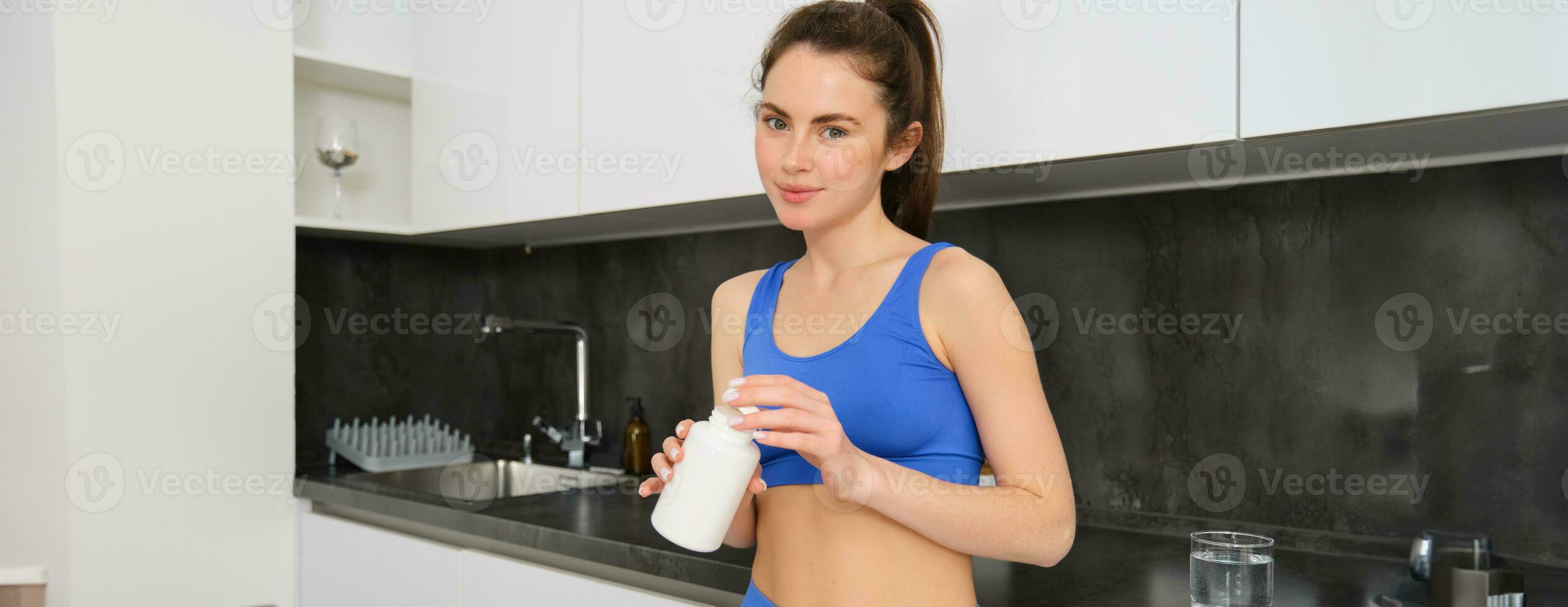 Portrait of brunette woman, wearing sport clothing, holding tablets, taking vitamins, dietary supplements buds for strong muscles, better workout results photo