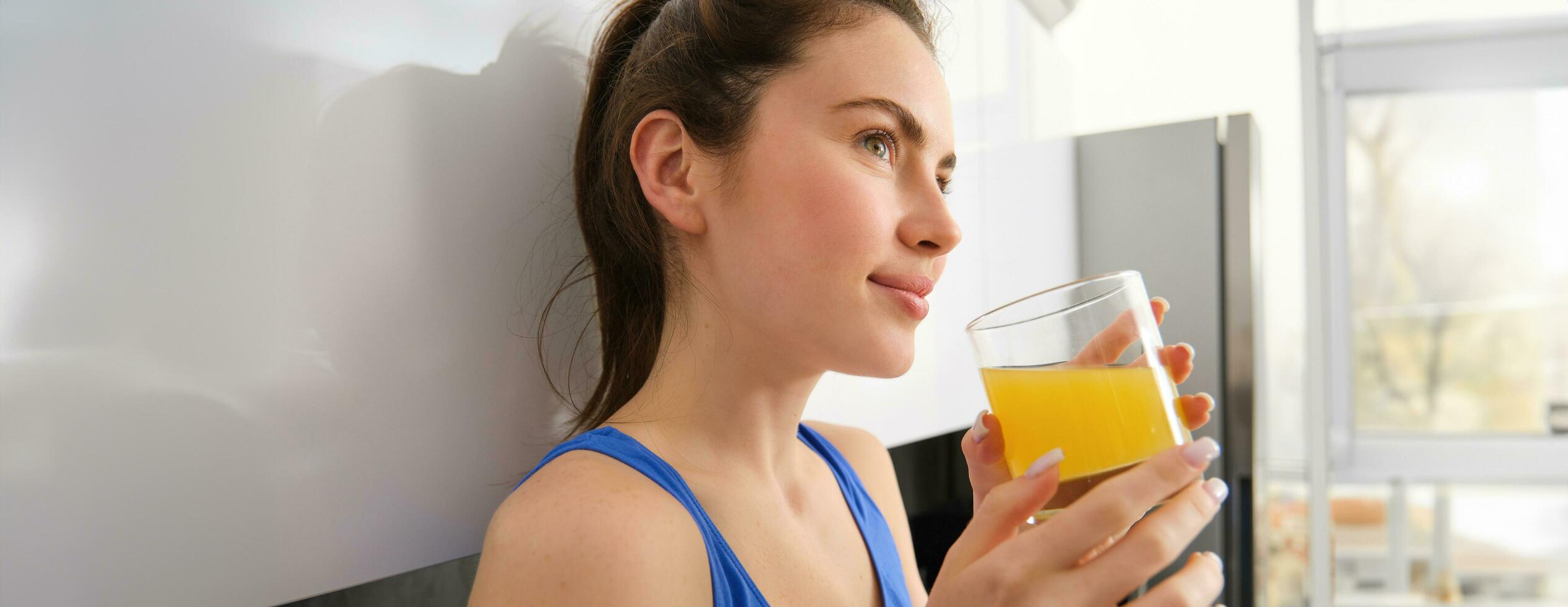 Close up portrait of beautiful young woman, looking away, drinking orange juice, fresh in glass, standing in kitchen, wearing workout clothes photo