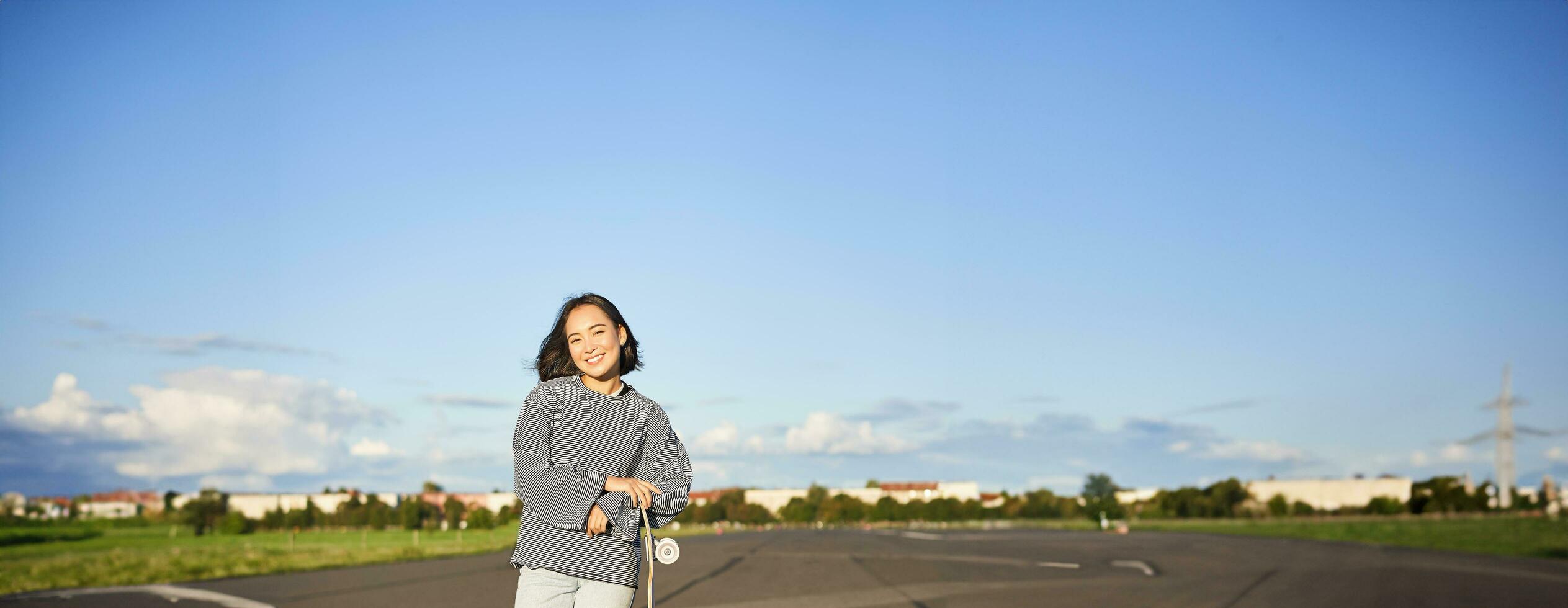 Vertical portrait of smiling asian woman standing on road with longboard, skateboarding on long cruiser, posing on empty road on sunny day photo