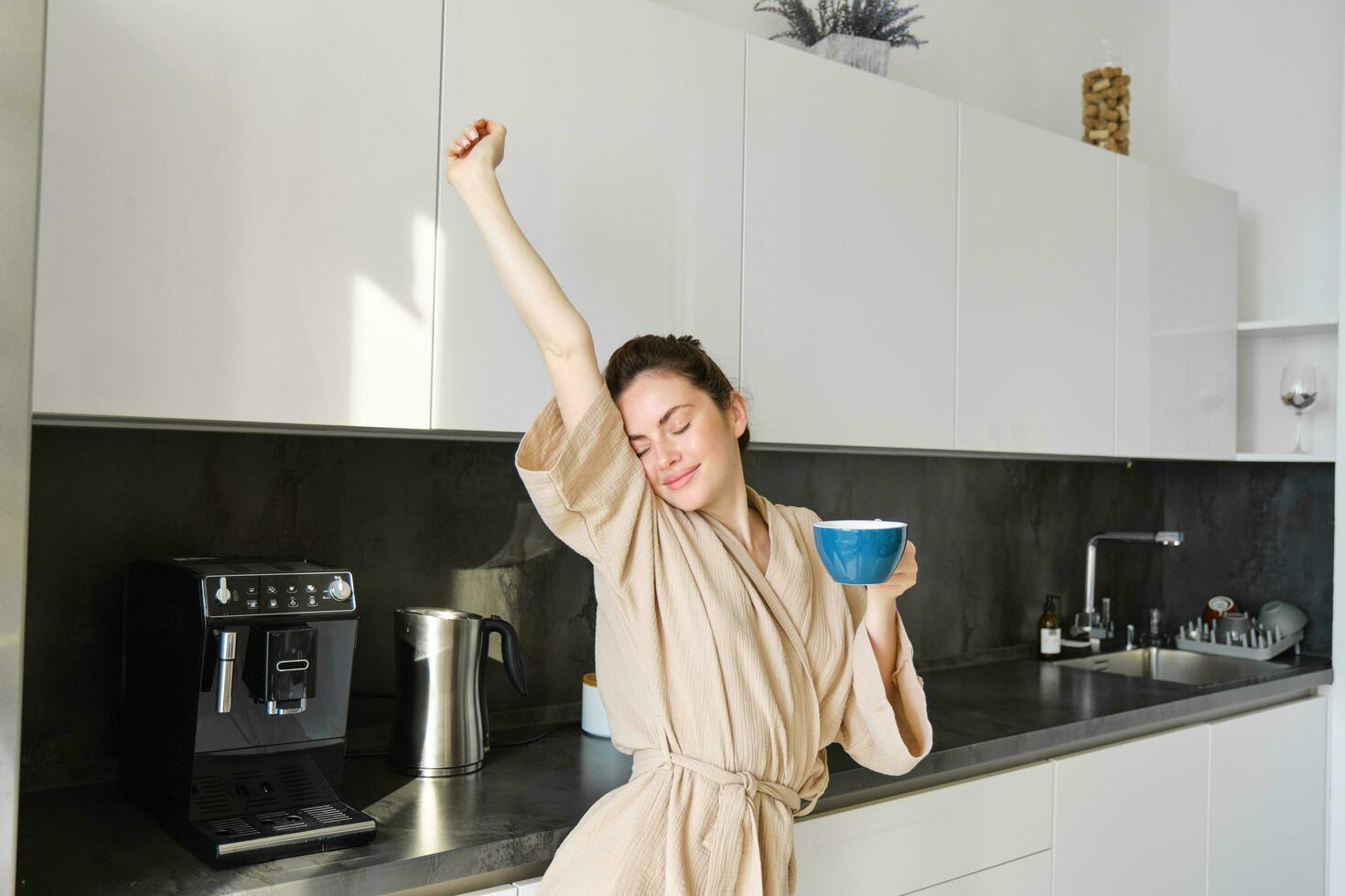 Portrait of happy girl dancing with coffee in the kitchen, wearing bathrobe, enjoying her morning routine photo