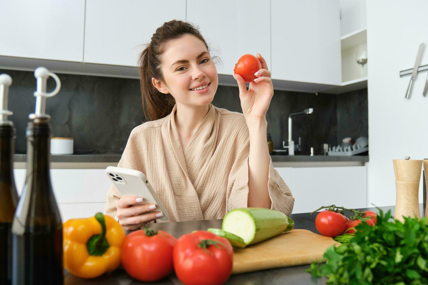 Young woman orders groceries on mobile app. Girl in bathrobe sits in the kitchen with vegetables, looking for recipe to cook dinner, using smartphone application photo
