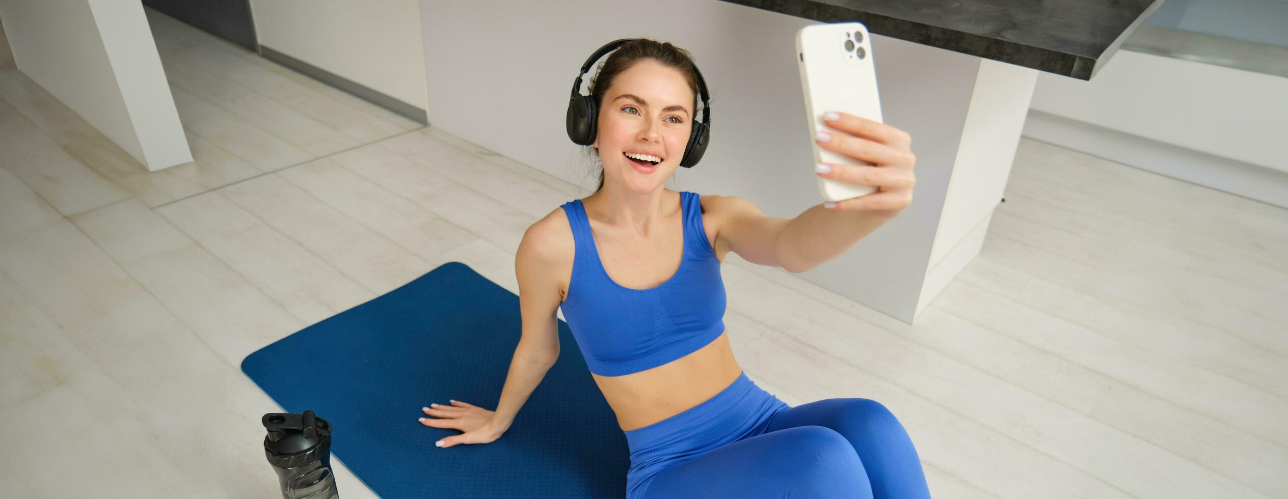 Portrait of fitness woman in wireless headphones, sits on fitness mat, doing selfies and photos during training exercises, working out at home