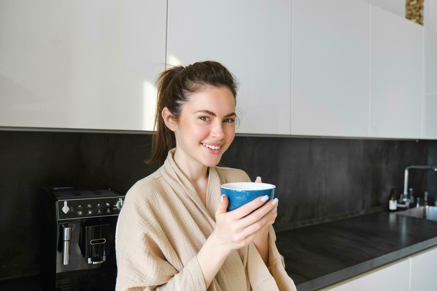 Portrait of carefree smiling woman drinking coffee, standing in kitchen with delighted, pleased face expression, waking up in the morning and having cappuccino photo