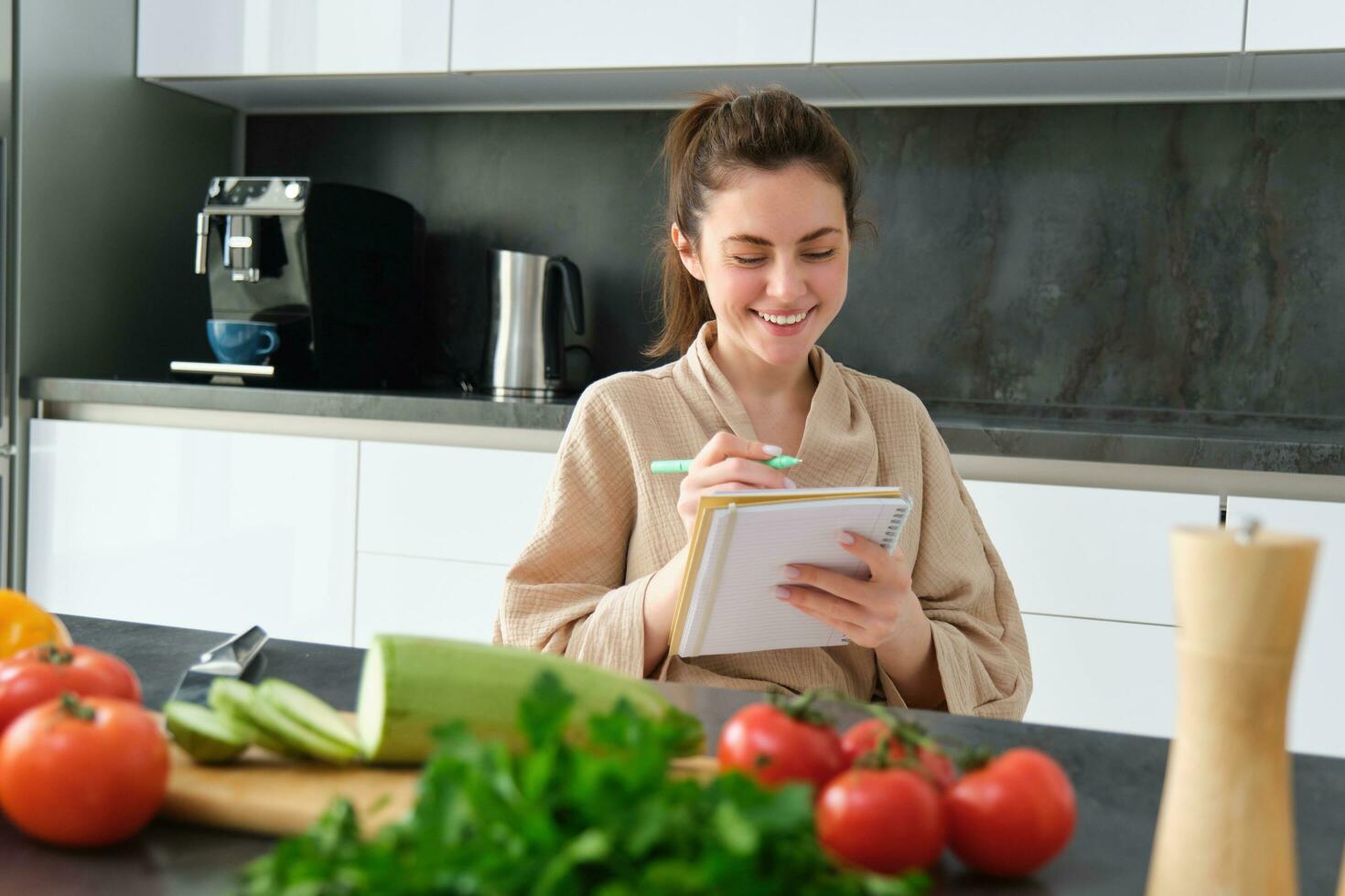 Portrait of beautiful, smiling young woman making list of meals, writing down recipe, sitting in the kitchen with vegetables, doing house errands photo