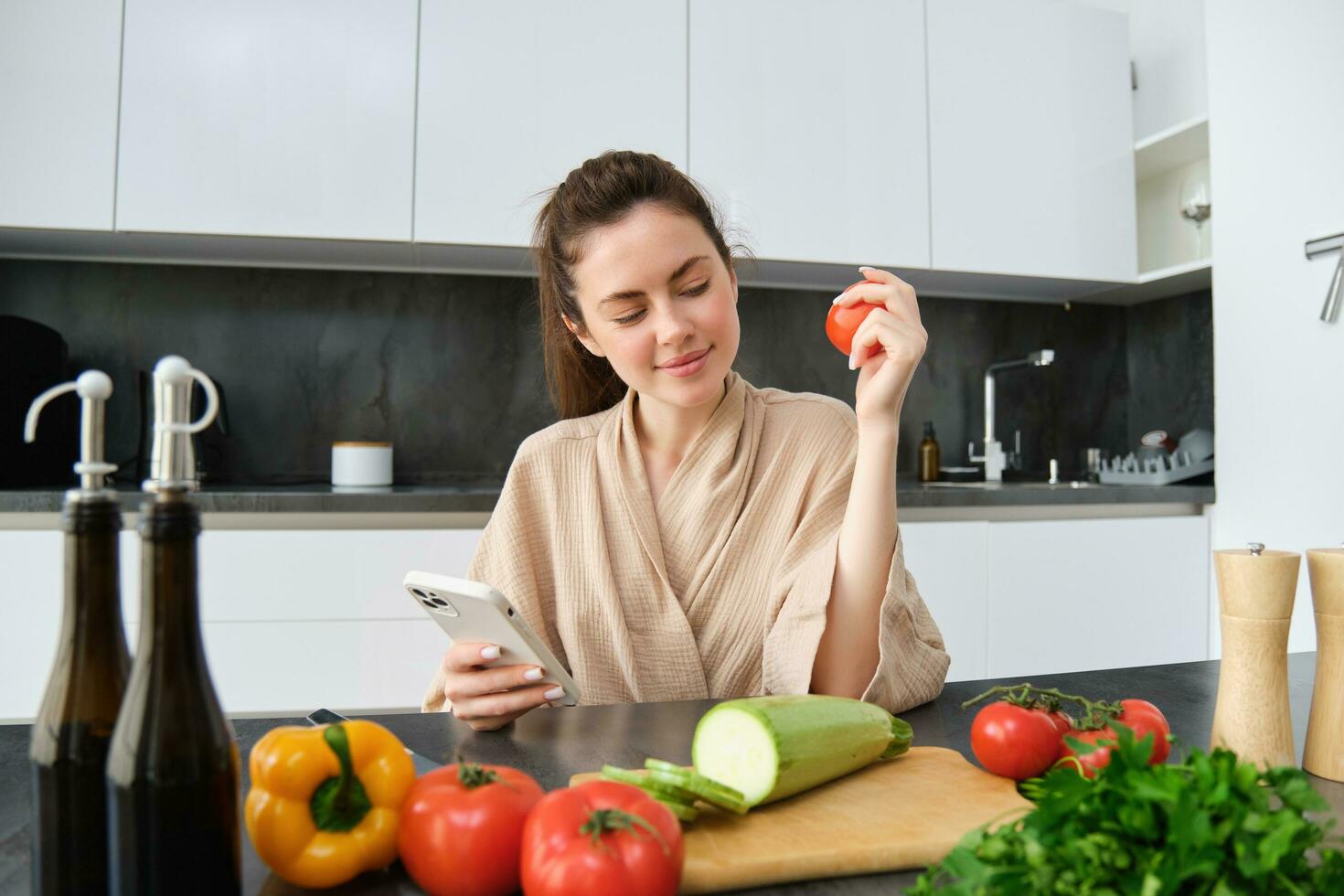 Image of young woman, chef on kitchen, cooking dinner at home, sitting near chopping board with vegetables and ingredients, using smartphone app to find recipe photo