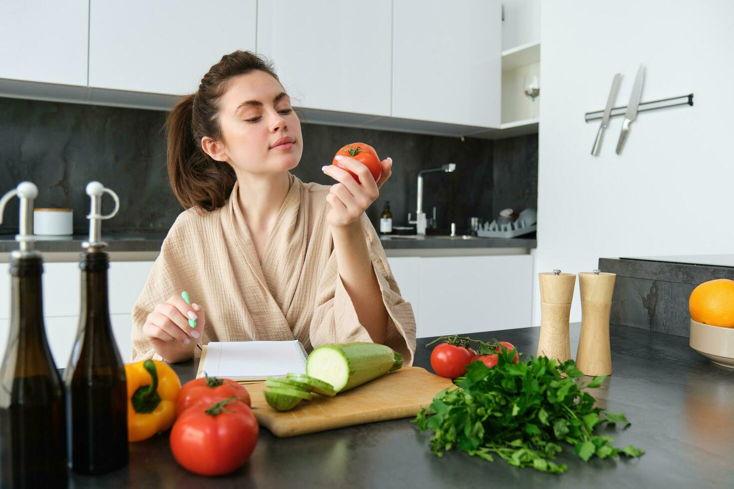 Portrait of beautiful smiling woman, writing her healthy menu, eating tomato while cooking, making grocery list, sitting in the kitchen photo