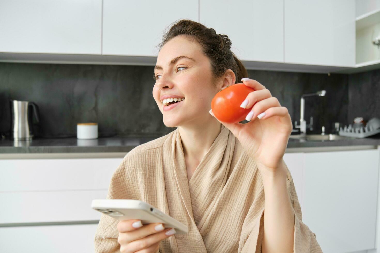 Young woman thinks what to cook, sits in the kitchen with smartphone and tomato in hands, looks aside and smiles, searches recipes on mobile app, orders groceries photo