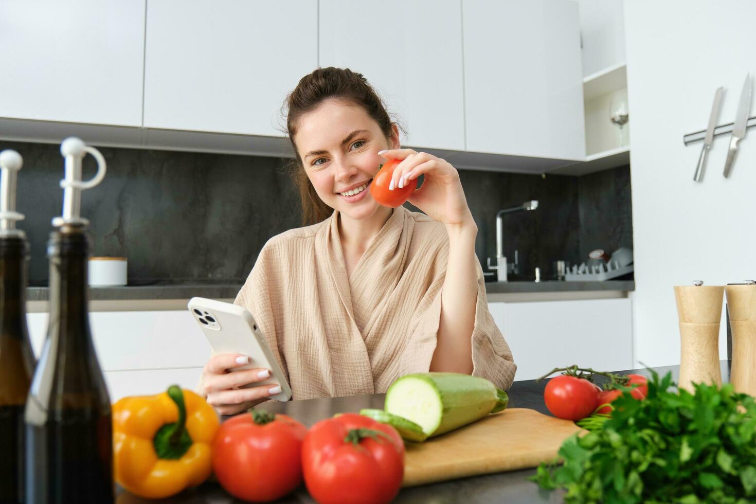 Portrait of young woman ordering groceries on smartphone app, holding tomato, sitting near chopping board with vegetables. Girl looking for recipe online, using mobile phone photo