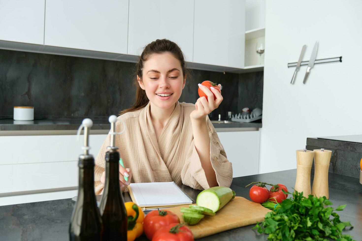 Portrait of beautiful smiling woman, writing her healthy menu, eating tomato while cooking, making grocery list, sitting in the kitchen photo