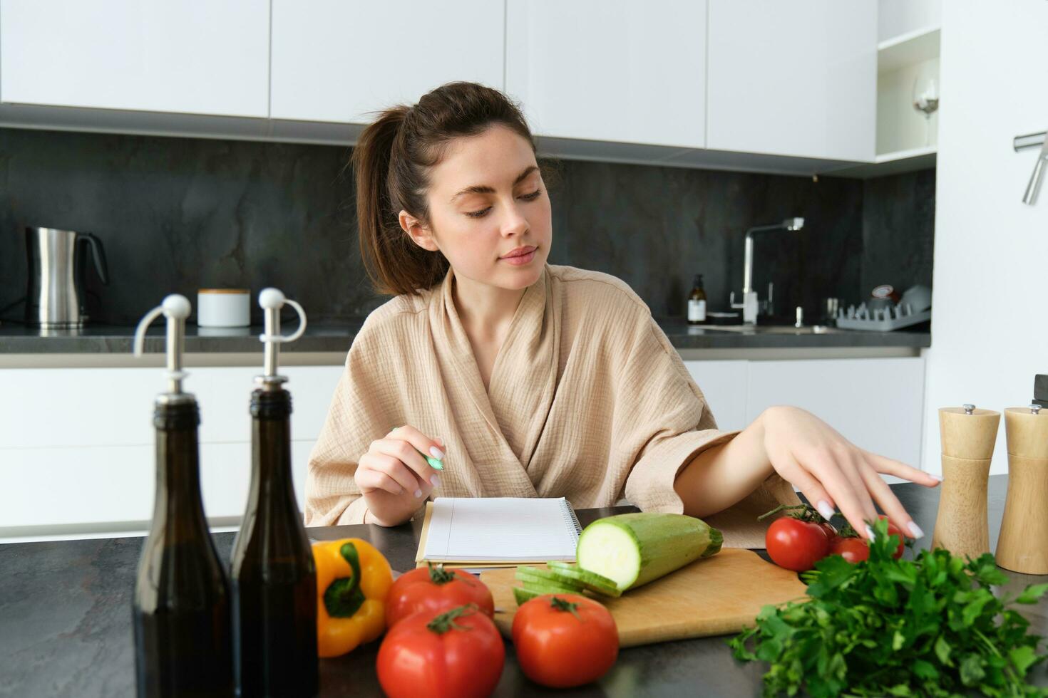 Portrait of woman writing down list of groceries, making notes in recipe, sitting in kitchen near vegetables, preparing dinner menu photo