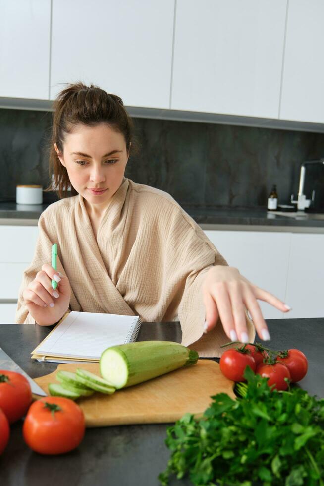 Portrait of woman writing down list of groceries, making notes in recipe, sitting in kitchen near vegetables, preparing dinner menu photo