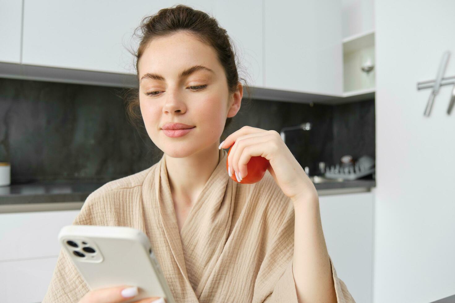 Close up portrait of beautiful smiling woman, holding fresh tomato, sitting in kitchen with smartphone, orders vegetables online, using application to buy groceries, using mobile phone photo