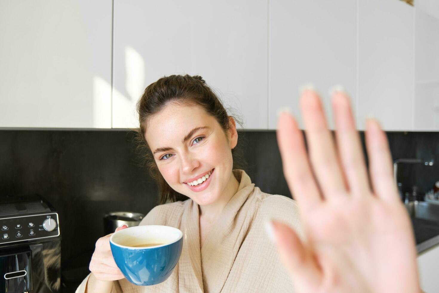 Portrait of cute young woman, covers herself from camera, laughing and flirting, drinking coffee in the morning, standing in the kitchen photo