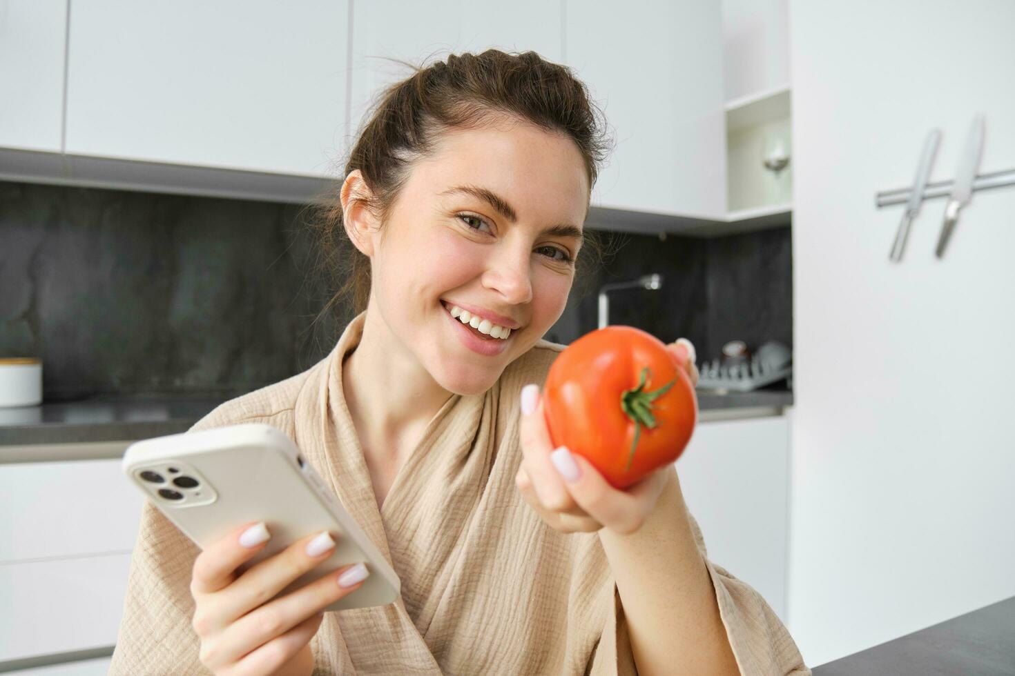 Image of smiling beautiful woman, sits in kitchen, holds smartphone and tomato, looks happy, orders groceries delivery to her home, using mobile phone app, buying online vegetables for her recipe photo