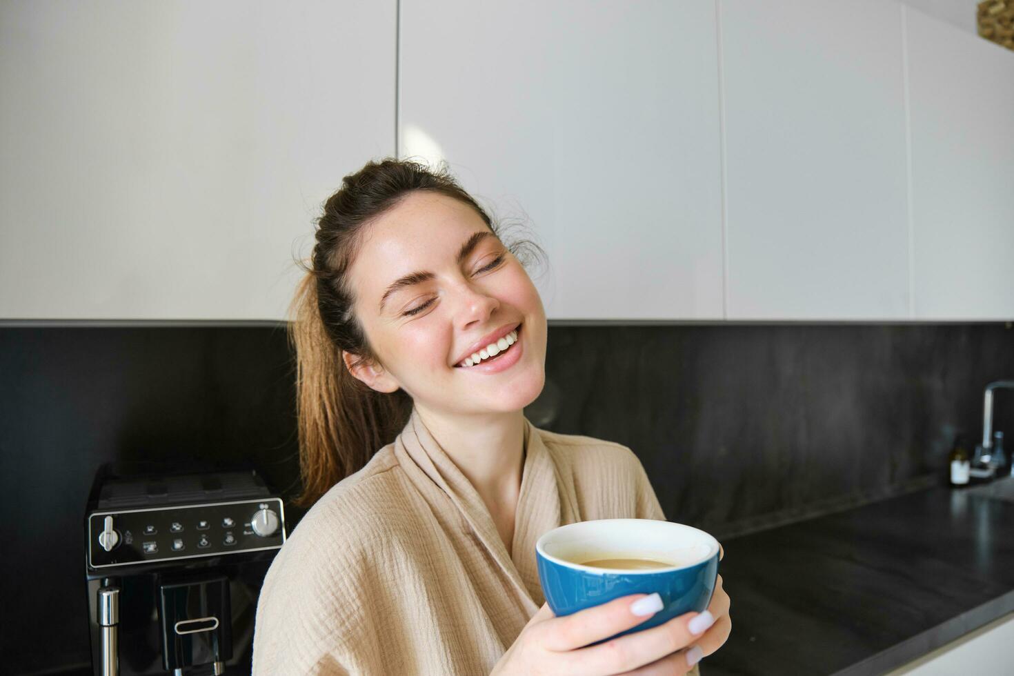 Portrait of carefree smiling woman drinking coffee, standing in kitchen with delighted, pleased face expression, waking up in the morning and having cappuccino photo
