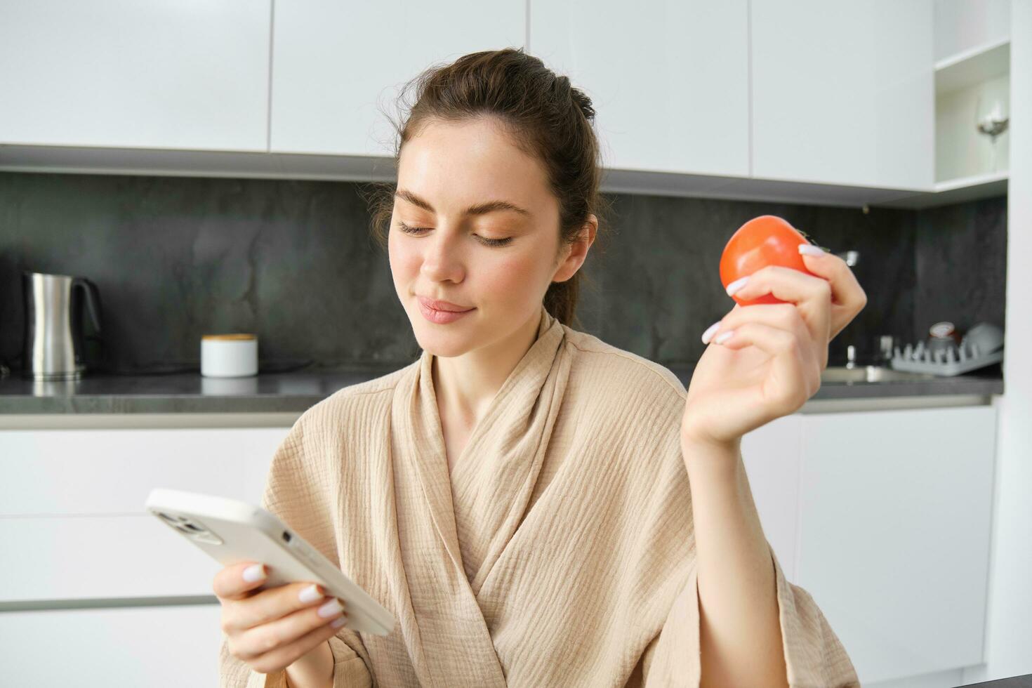 Close up portrait of beautiful smiling woman, holding fresh tomato, sitting in kitchen with smartphone, orders vegetables online, using application to buy groceries, using mobile phone photo