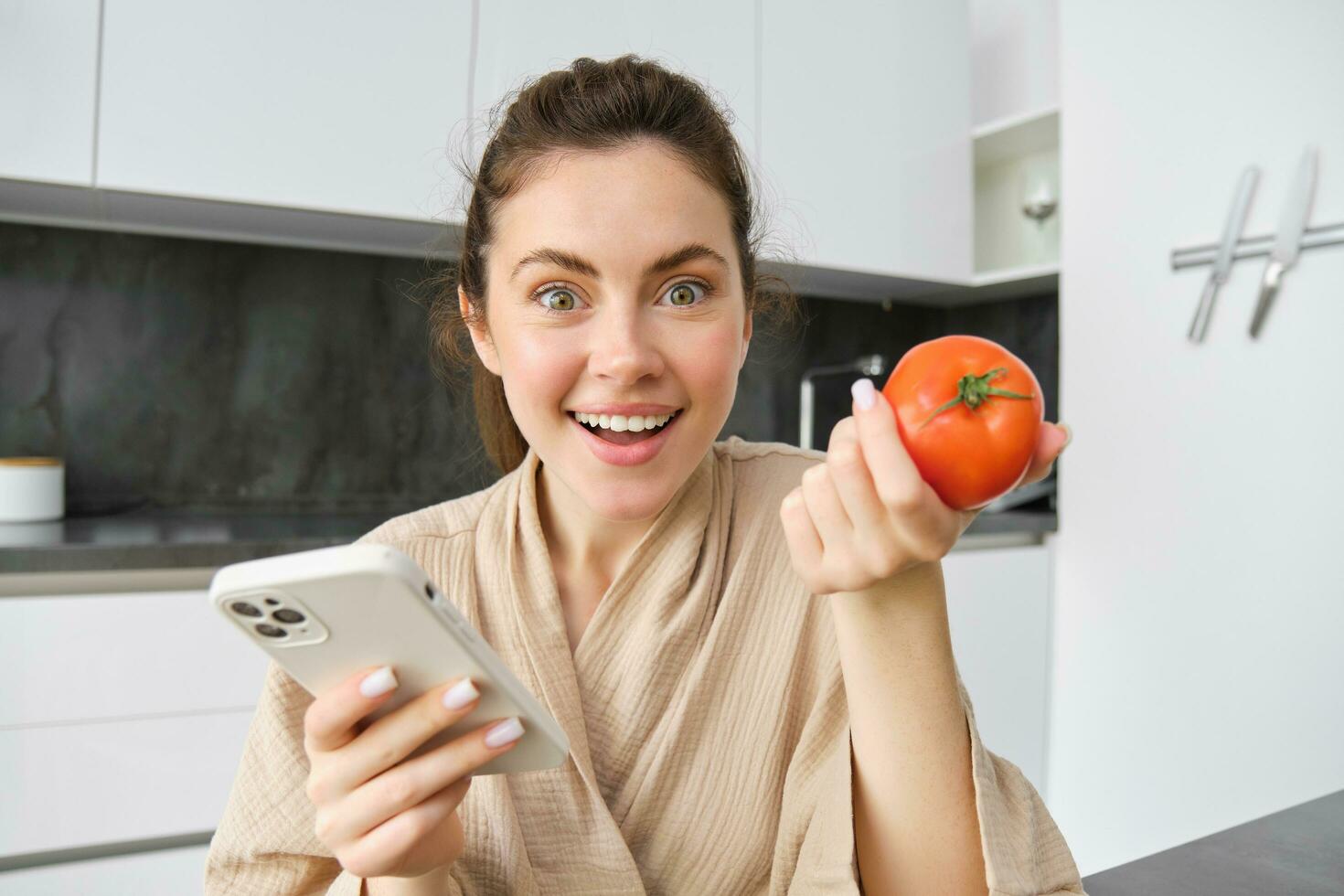 Close up portrait of beautiful young woman in the kitchen, holding tomato and smartphone, buys fresh vegetables online, orders groceries delivery on mobile phone application photo