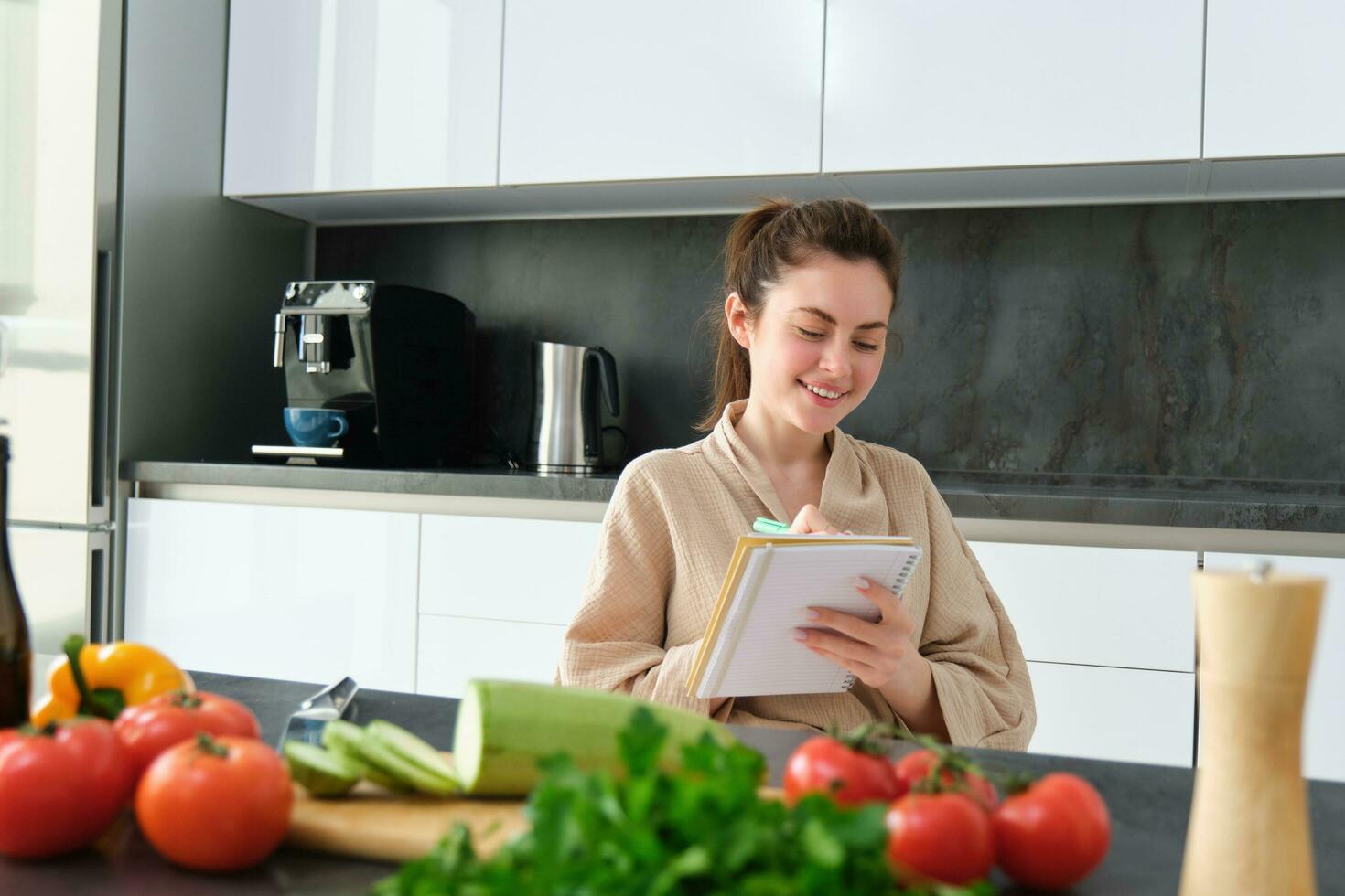 retrato de hermoso, sonriente joven mujer haciendo lista de comidas, escritura abajo receta, sentado en el cocina con verduras, haciendo casa Diligencias foto