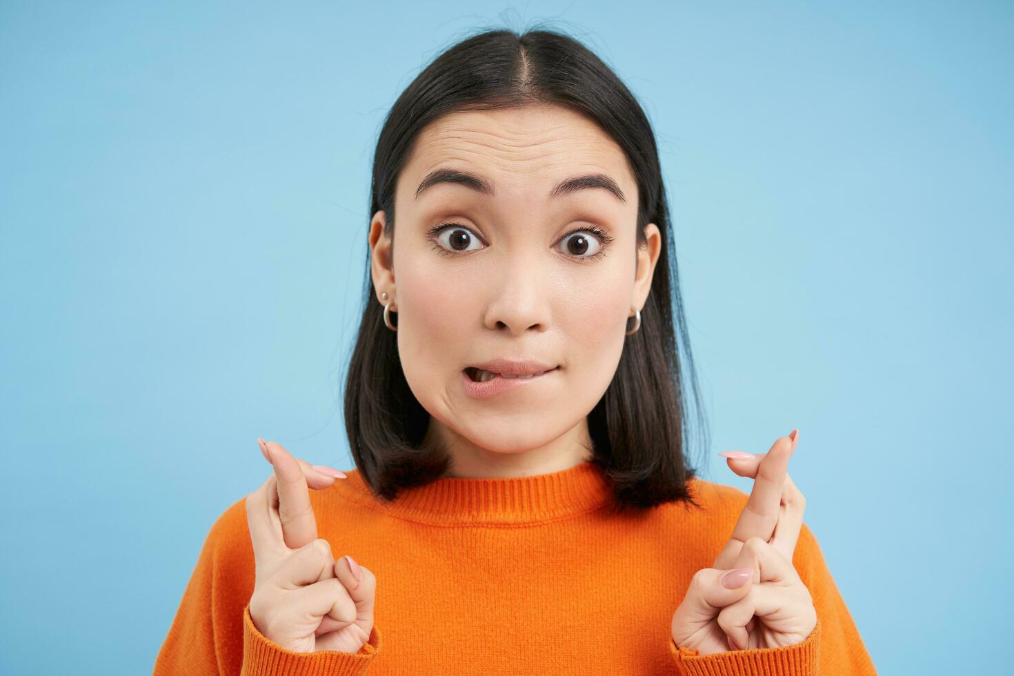 Close up of hopeful asian girl, makes wish, cross fingers, stands over blue background photo