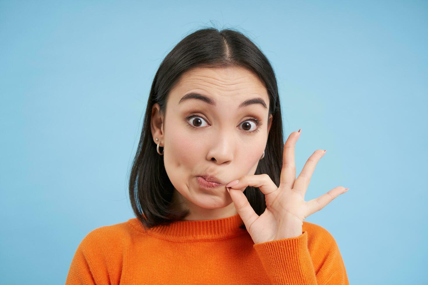 Gossips. Close up of cute asian girl zips mouth, locks lips on seal, promise to keep secret, stands over blue background photo